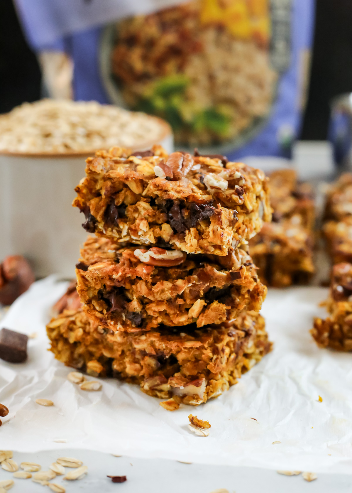 A stack of pumpkin oats bars with a dense, crumbly texture are stacked on a piece of white parchment paper on a kitchen countertop, with additional pumpkin bars scattered in the background