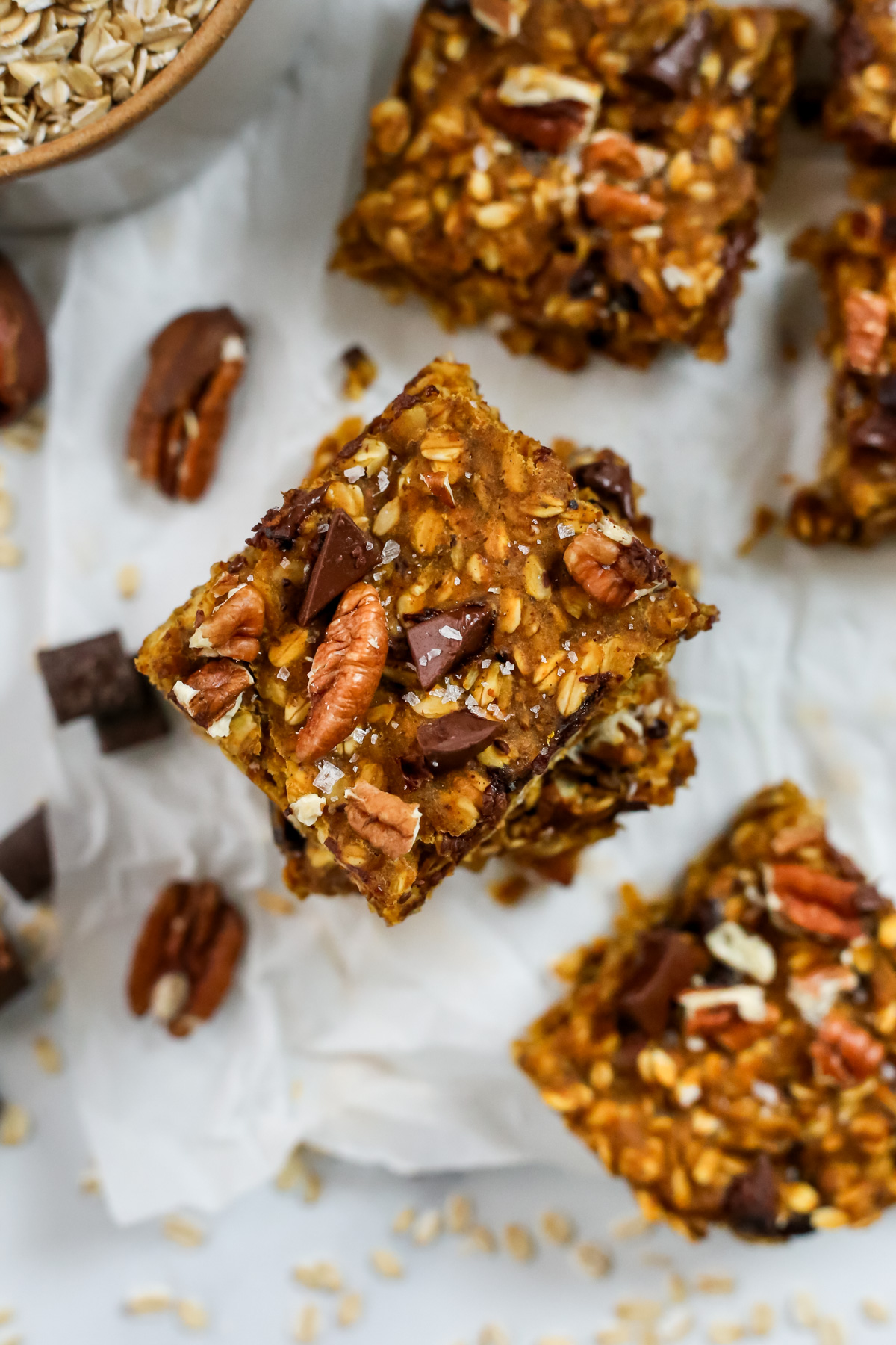 Overhead view of a stack of pumpkin oats bars topped with pecan pieces and dark chocolate chunks, sprinkled with flaky sea salt and displayed on a piece of white parchment paper