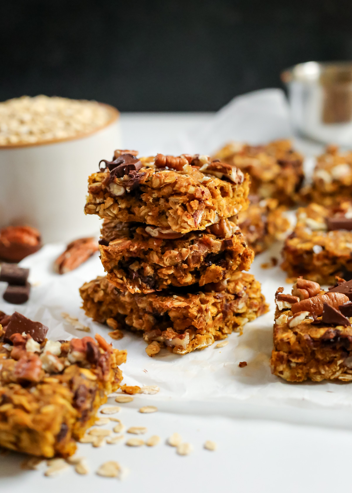 A stack of three pumpkin oat bars is stacked in front of a dark black background and displayed among other bars as if they were just sliced. The texture is dense and crumbly with visible chunks of chocolate and pecans mixed throughout