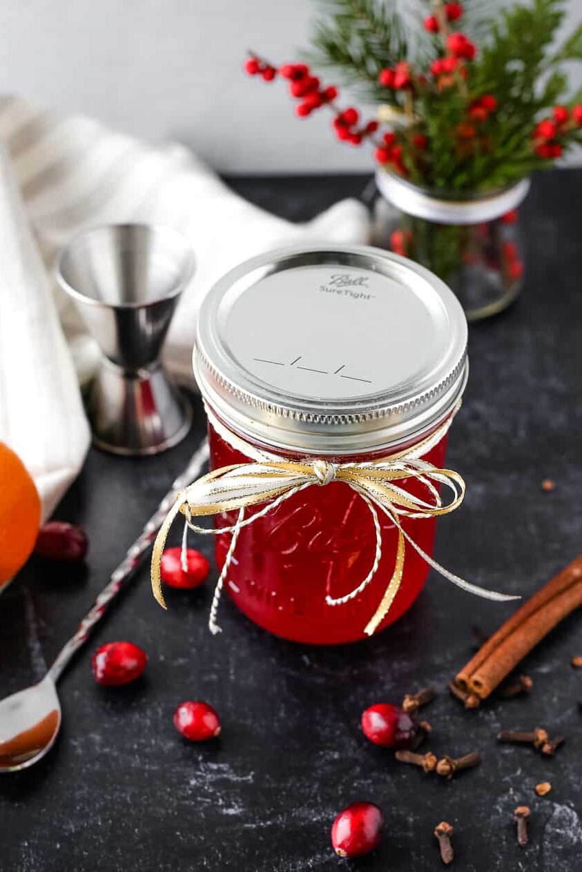 Angled view of a glass mason jar with the lid fastened displayed in a holiday scene. The jar is full of a vibrant red homemade cranberry simple syrup and displayed as a DIY gift