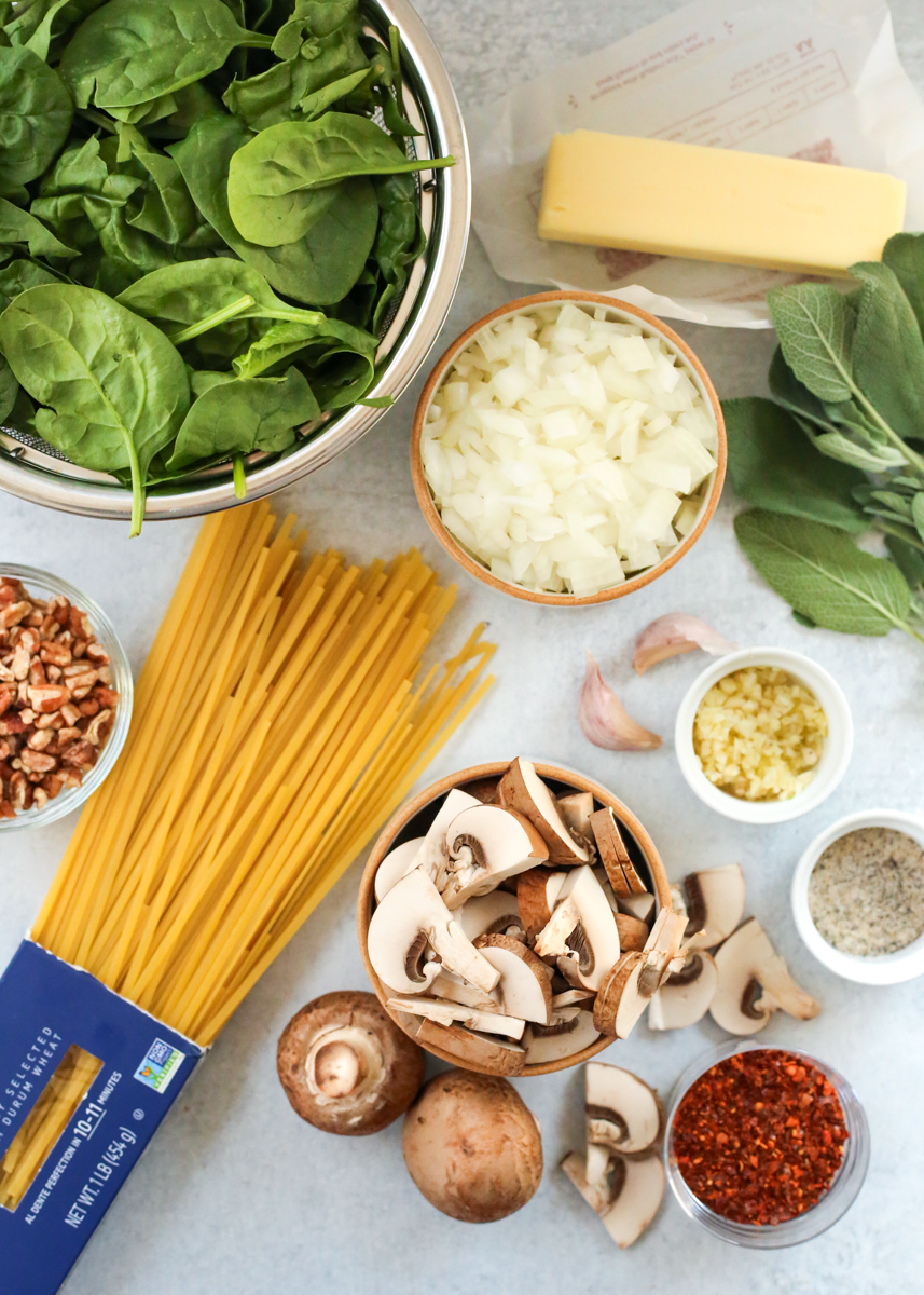 Overhead view of the ingredients needed to make a brown butter mushroom pasta recipe, including uncooked linguine pasta, fresh spinach, diced onions, a stick of butter, fresh sage leaves, garlic cloves, sliced mushrooms, red pepper flakes, and chopped pecans, displayed in various prep bowls or small ramekins