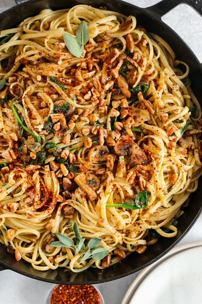 A close overhead view into a skillet full of cooked linguine pasta, topped with chopped pecans, sautéed mushrooms, chopped pecans, cooked spinach, fresh sage, and red pepper flakes