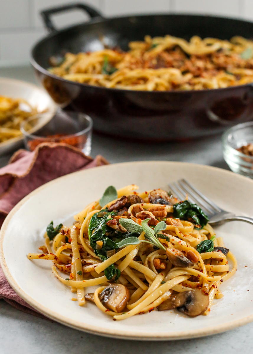 A styled plate with brown butter mushroom pasta is served in front of a skillet with the rest of the pasta recipe