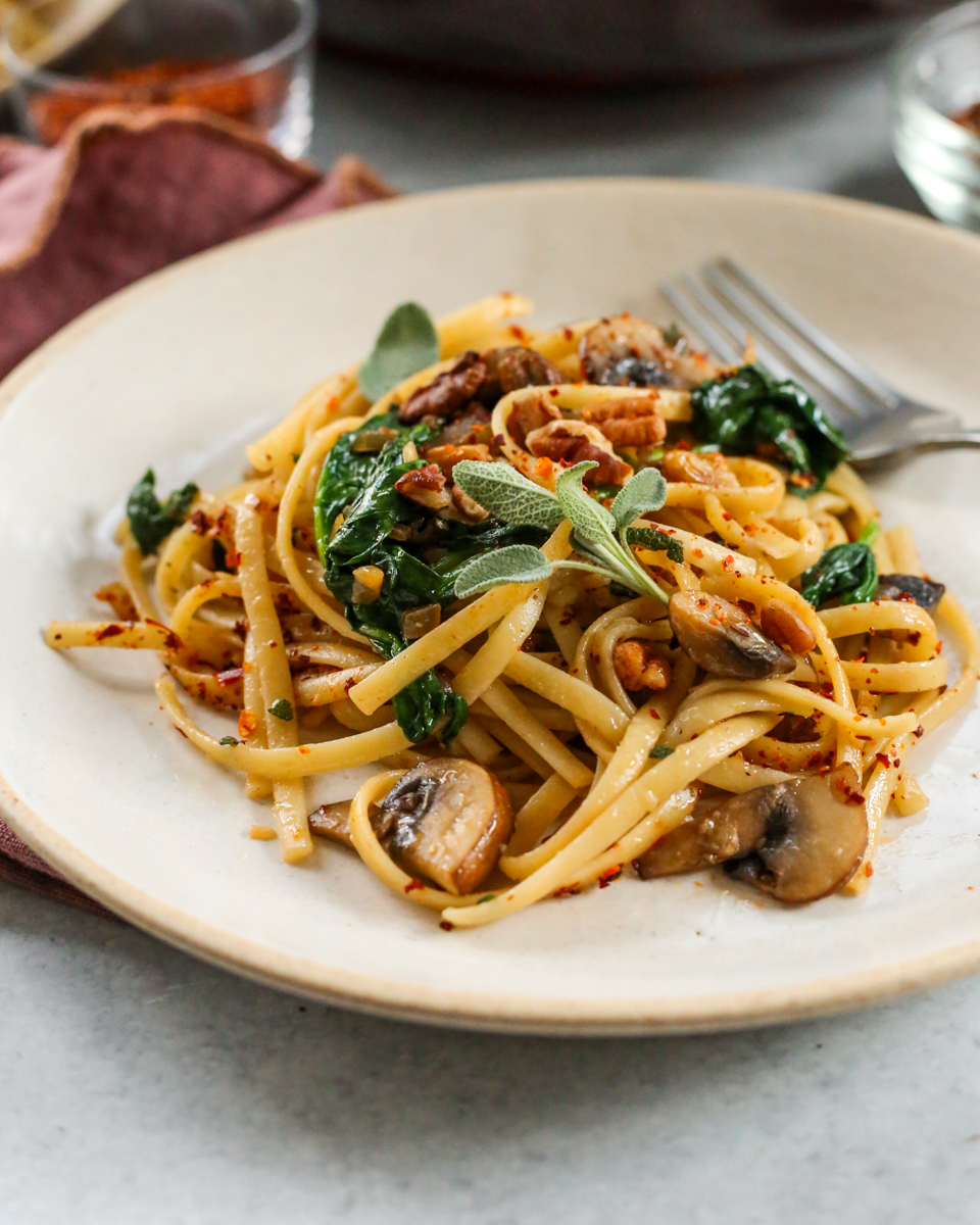 An angled view at a served portion of brown butter mushroom pasta, garnished with fresh sage and red pepper flakes, with visible pieces of cooked mushroom and spinach mixed throughout