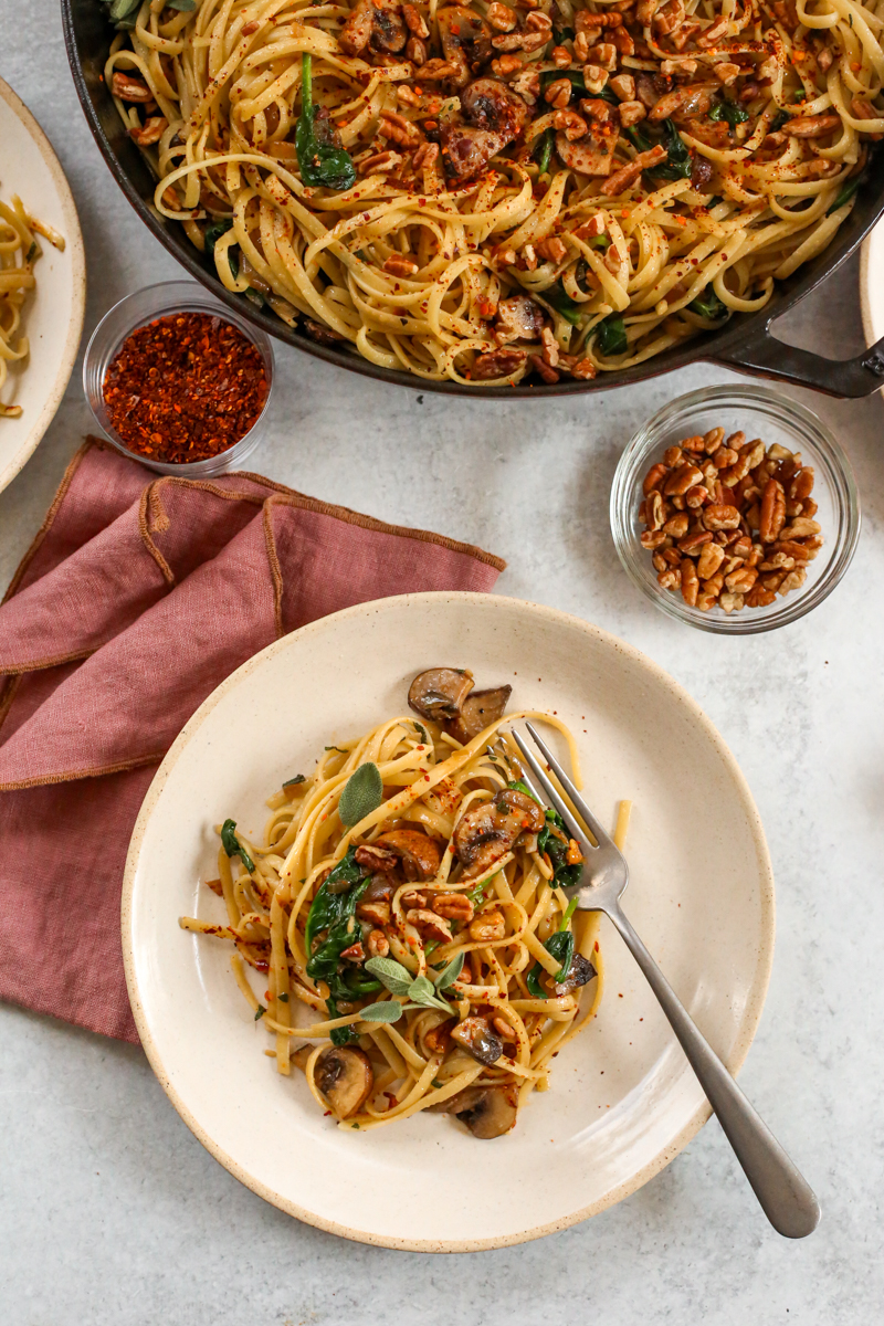 A kitchen scene with a serving plate with brown butter mushroom pasta alongside the serving bowl, with additional garnishes placed nearby and silverware nearby as if ready to take a bite