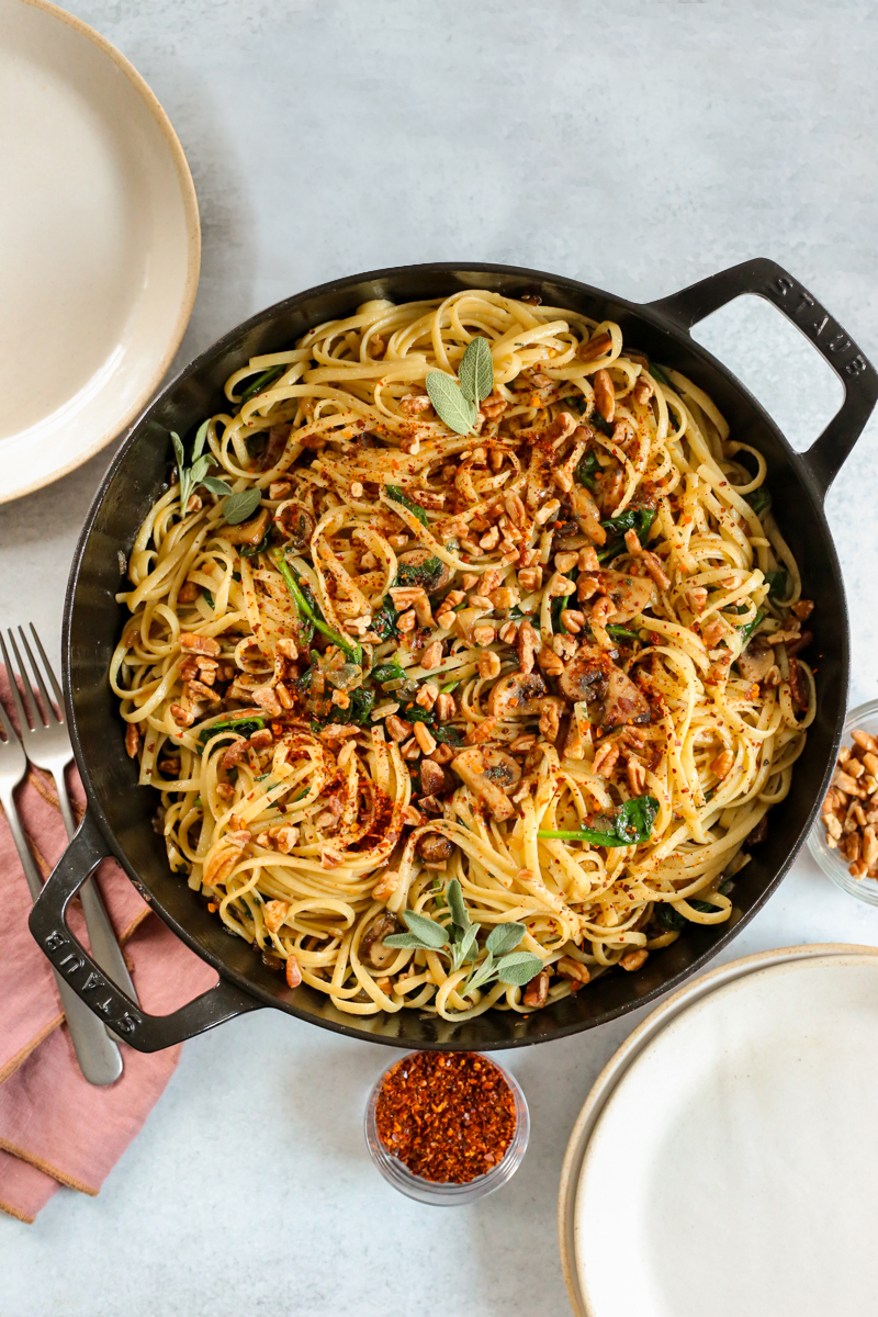 Overhead view into a large braising pan filled with cooked linguine pasta with a brown butter sauce, topped with pecans, fresh sage, and red pepper flakes, with serving plates and silverware arranged nearby on a kitchen countertop
