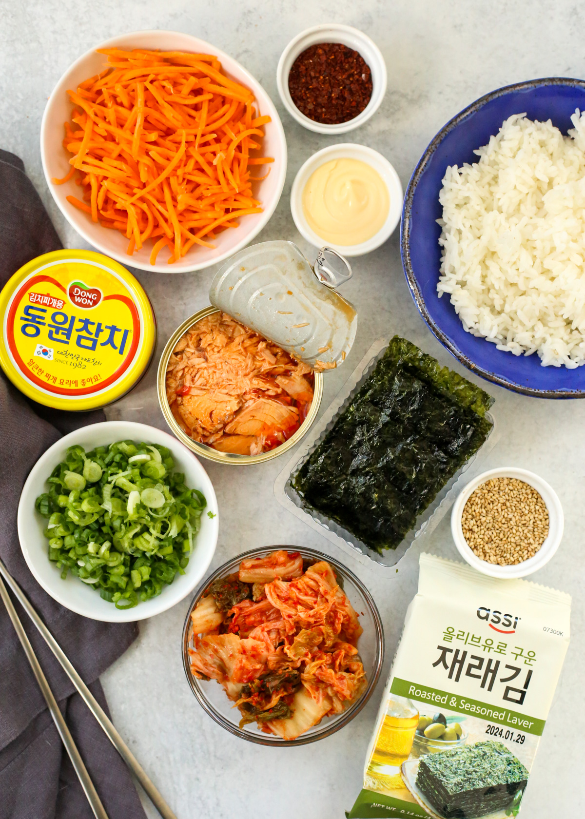 Flatlay of all the ingredients needed to a Korean tuna rice bowl, including canned tun, matchstick carrots, kewpie mayo, toasted sesame seeds, cooked white rice, a package of seaweed snack sheets, toasted sesame seeds, kimchi, and sliced green onions