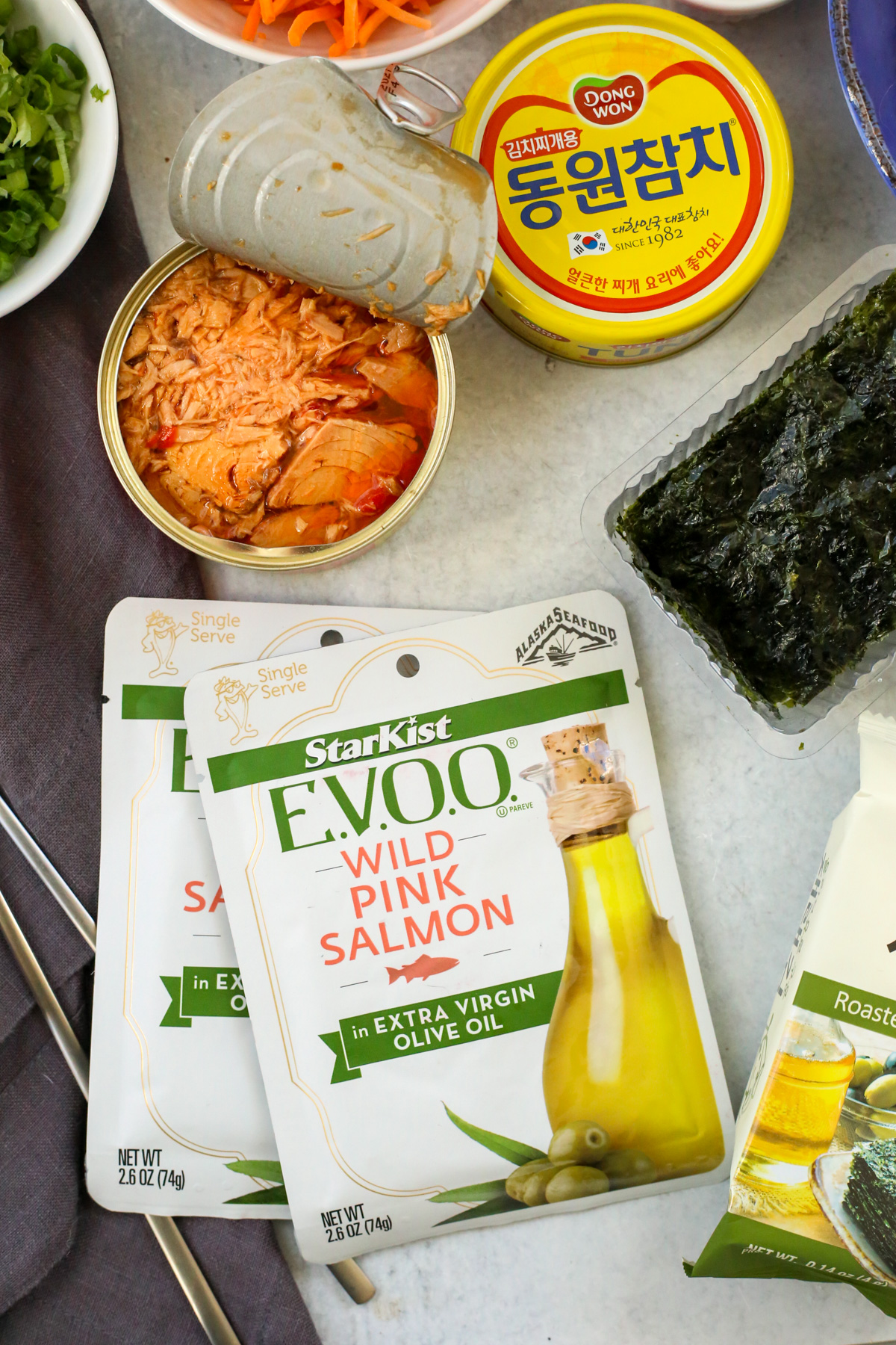 An open can of Korean style tuna is arranged next to two unopened pouches of Starkest Salmon packed in extra virgin olive oil, displayed on a kitchen countertop with a pair of silver chopsticks