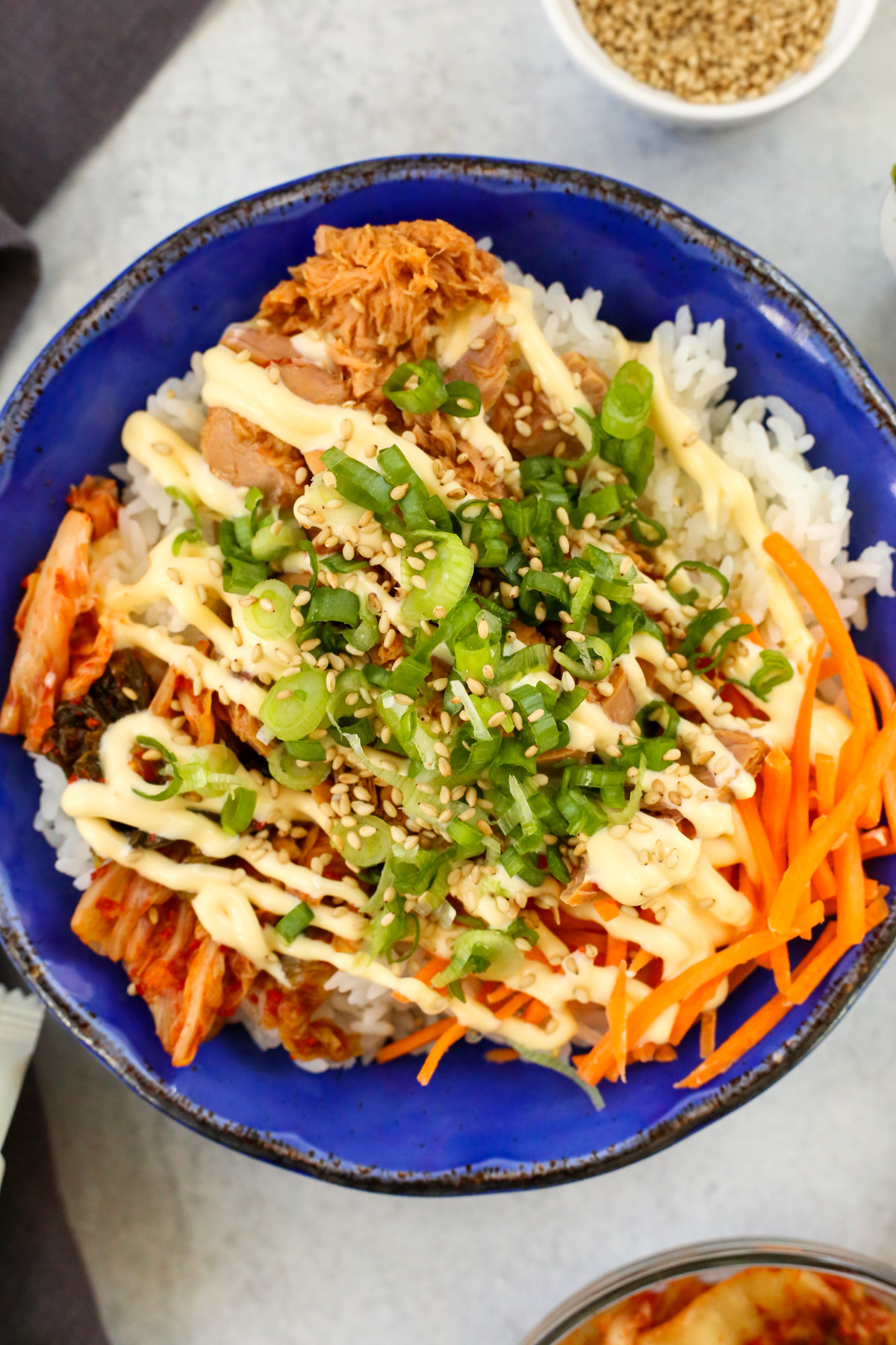 Closeup overhead view of a bowl of chamchi deopbap (Korean Tuna Rice Bowl) served in a bright blue glazed ceramic bowl, garnished heavily with kewpie mayo, sliced green onions, and toasted sesame seeds
