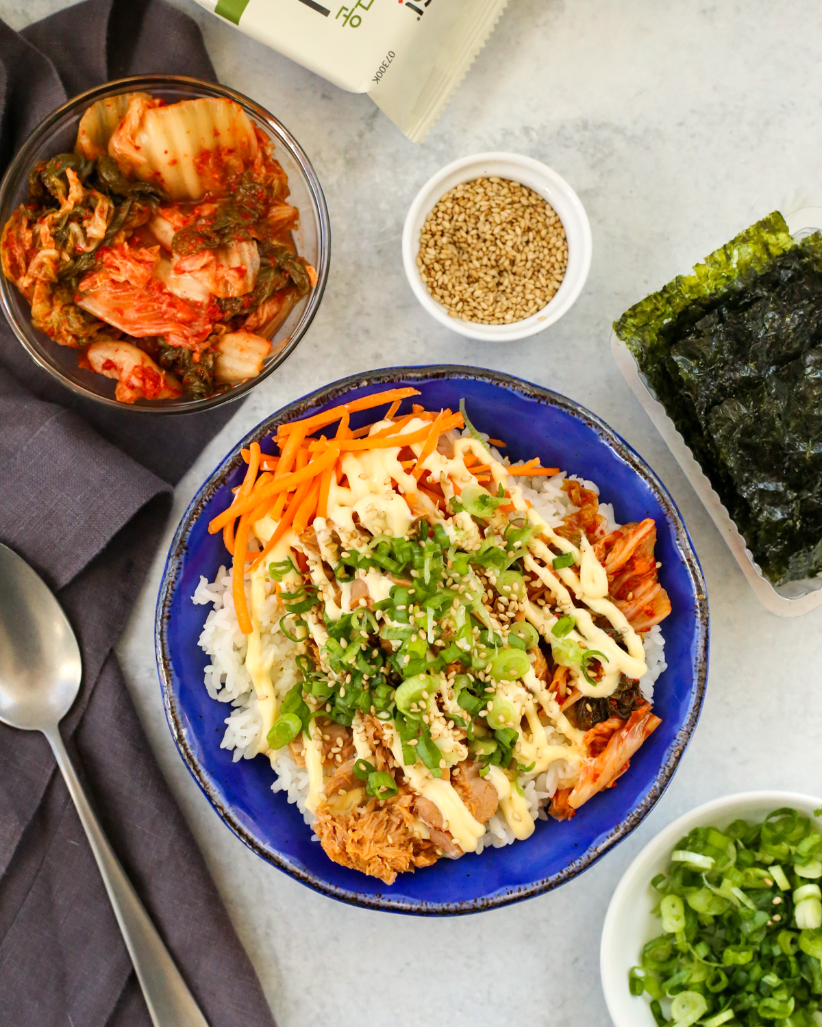 Overhead view of a glazed blue bowl filled with rice, canned tuna, kimchi, carrots, and garnished with kewpie mayo, sliced green onions, and toasted sesame seeds, with a silver spoon and small serving bowls arranged nearby