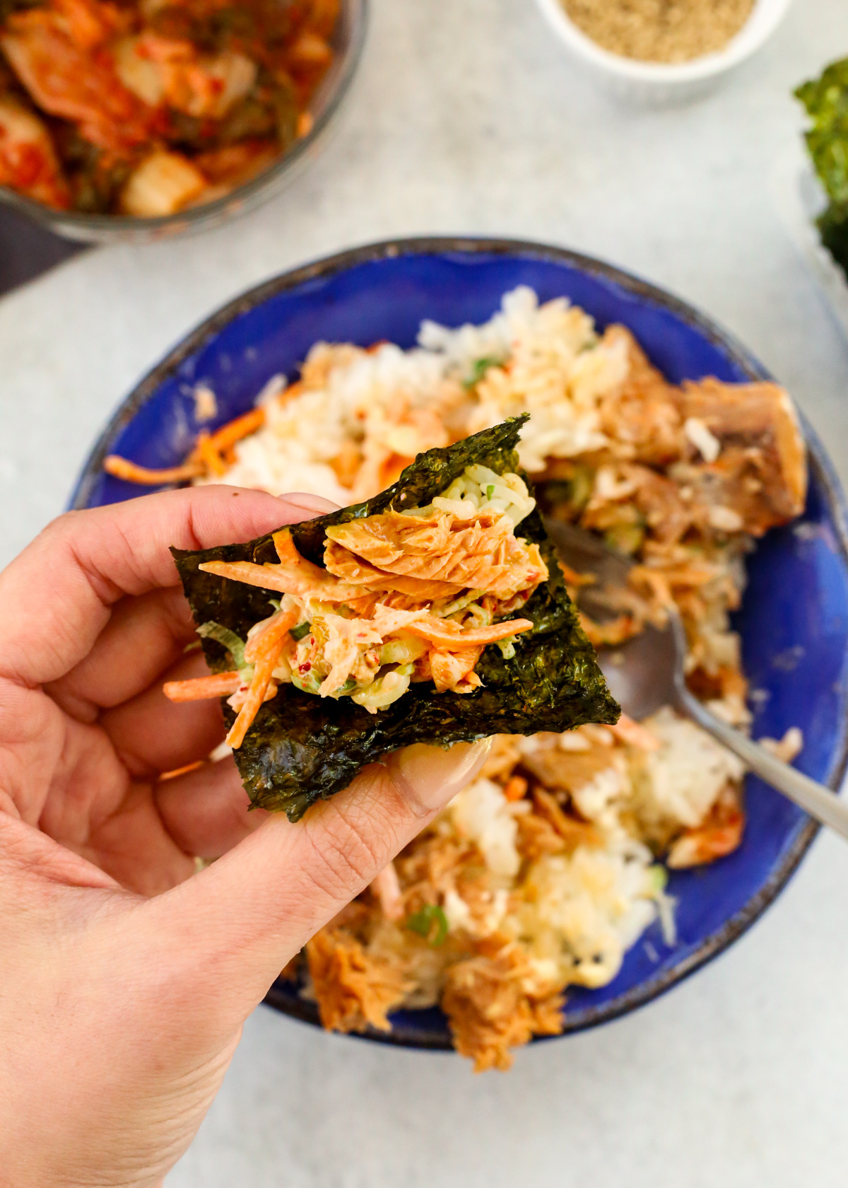 A woman's hand with manicured nails holds a bite of chamchi deopbap (Korean tuna rice bowl) wrapped in gim, a small sheet of dried and roasted seaweed, with the mixed bowl visible in the background