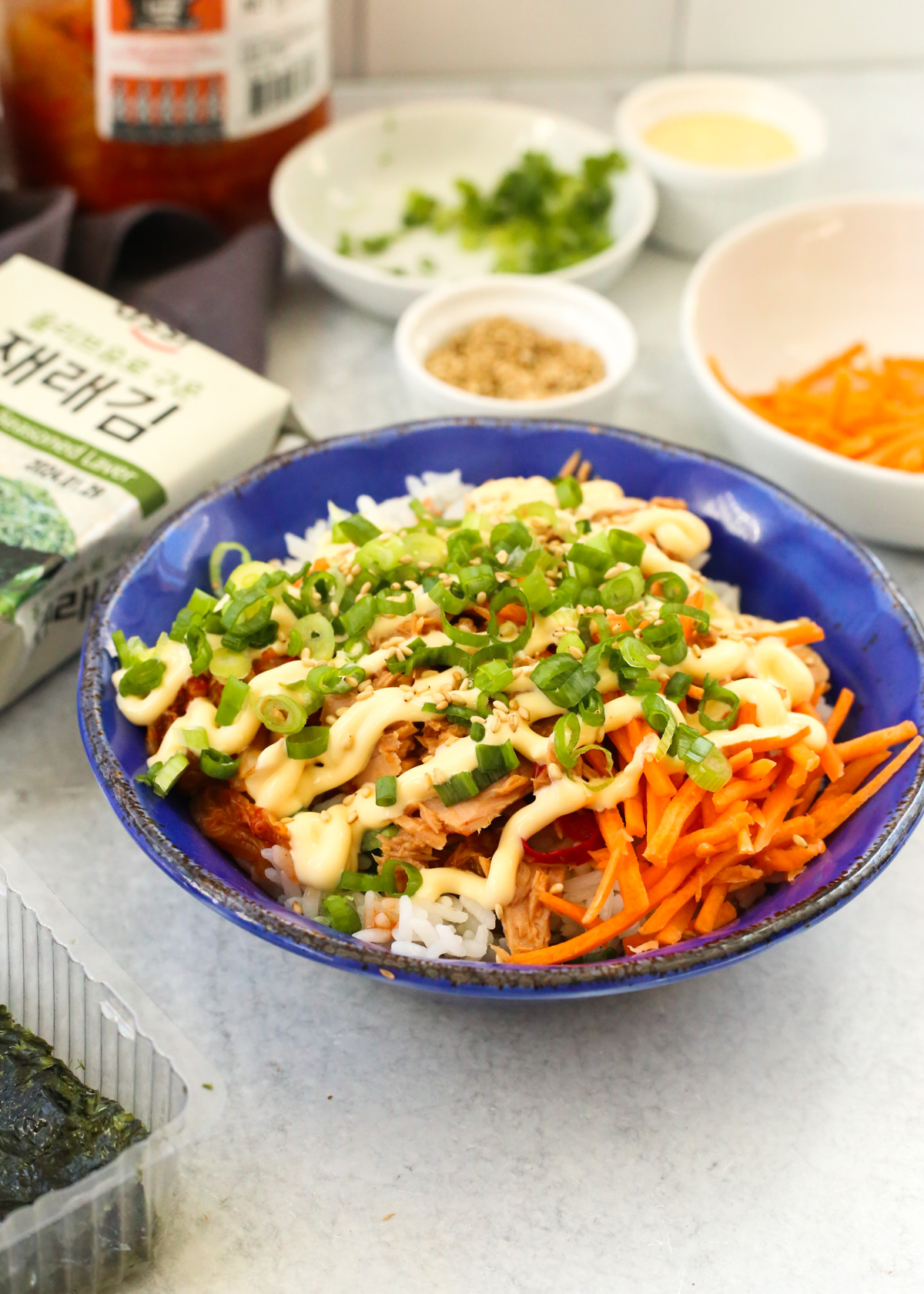 Styled bowl of chamchi deopbap (Korean Tuna Rice Bowl) in a glazed blue bowl, drizzled with mayo and garnished heavily with thinly sliced green onions and additional ingredients visible in the background