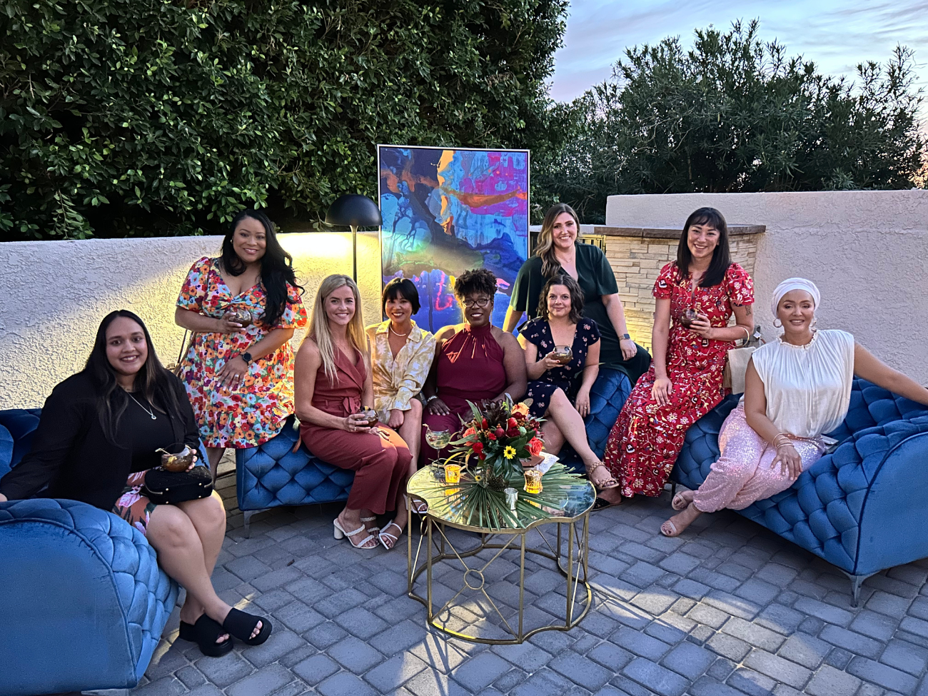 A groups of women in cocktail attire sit outdoors on a pool patio in sunset light. All are smiling at the camera and posing on various blue velvet furniture that appears posh and sophisticated