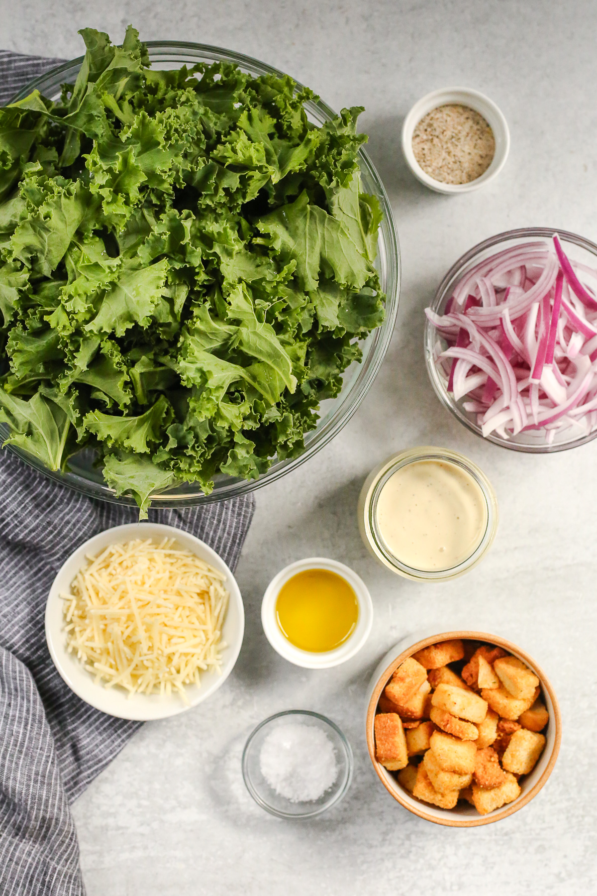 Overhead view of a kitchen countertop displaying the ingredients needed to make a kale caesar salad, including a large bowl of raw kale leaves, plus sliced red onion, Caesar salad dressing, parmesan cheese, and golden brown croutons in small prep bowls nearby.