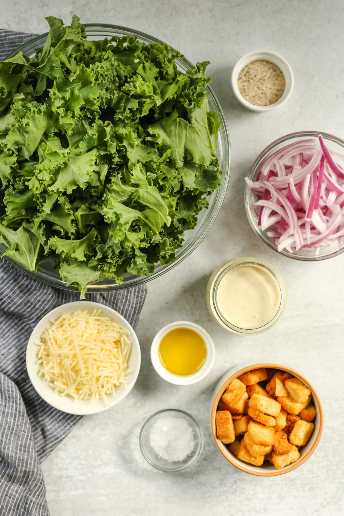 Overhead view of a kitchen countertop displaying the ingredients needed to make a kale caesar salad, including a large bowl of raw kale leaves, plus sliced red onion, Caesar salad dressing, parmesan cheese, and golden brown croutons in small prep bowls nearby