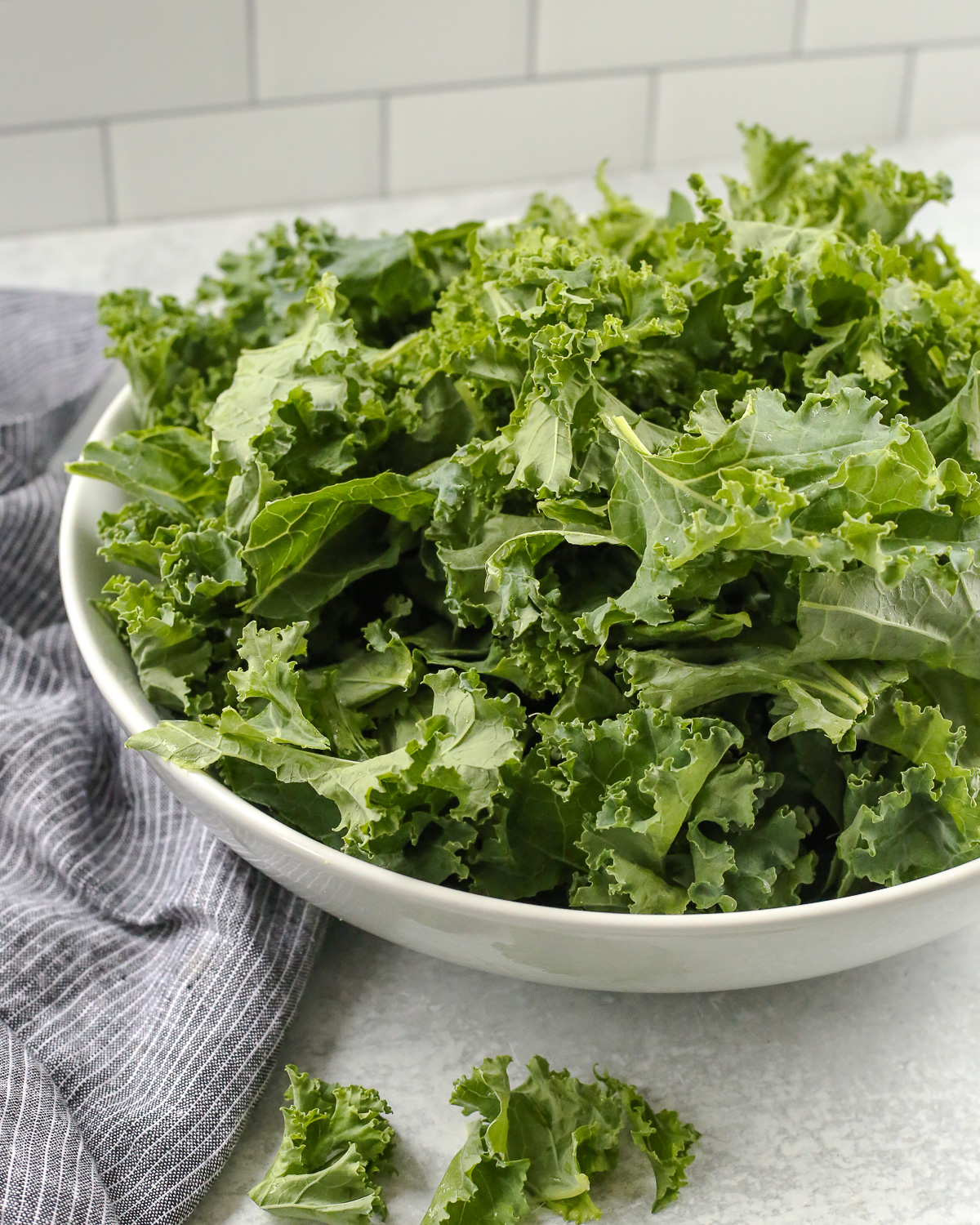 A white serving bowl full of raw kale, appearing fibrous and tough.
