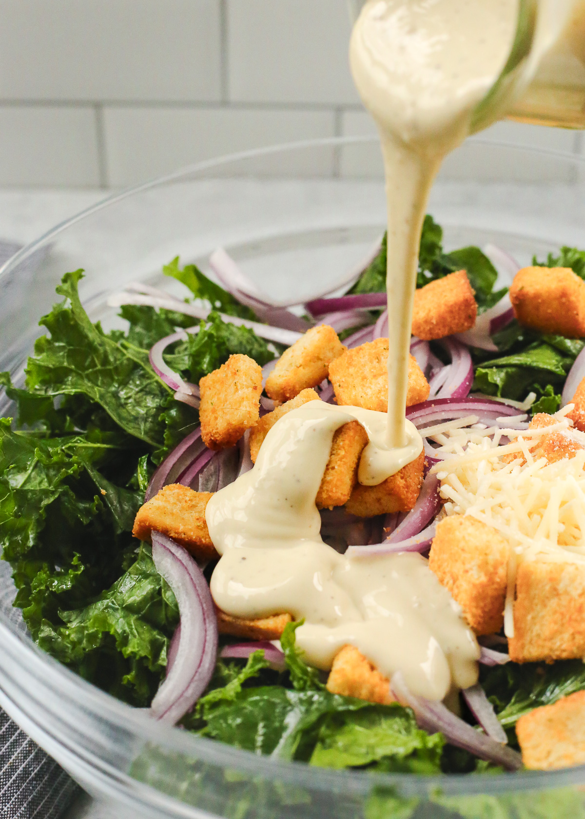 An angled view into a large mixing bowl filled with massaged kale, sliced red onions, parmesan cheese, and golden brown croutons, as creamy caesar salad dressing is poured into it from a small glass jar.