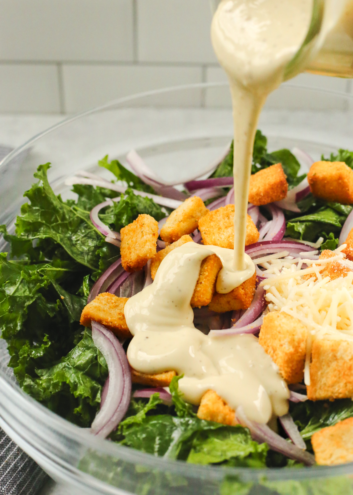 An angled view into a large mixing bowl filled with massaged kale, sliced red onions, parmesan cheese, and golden brown croutons, as creamy caesar salad dressing is poured into it from a small glass jar