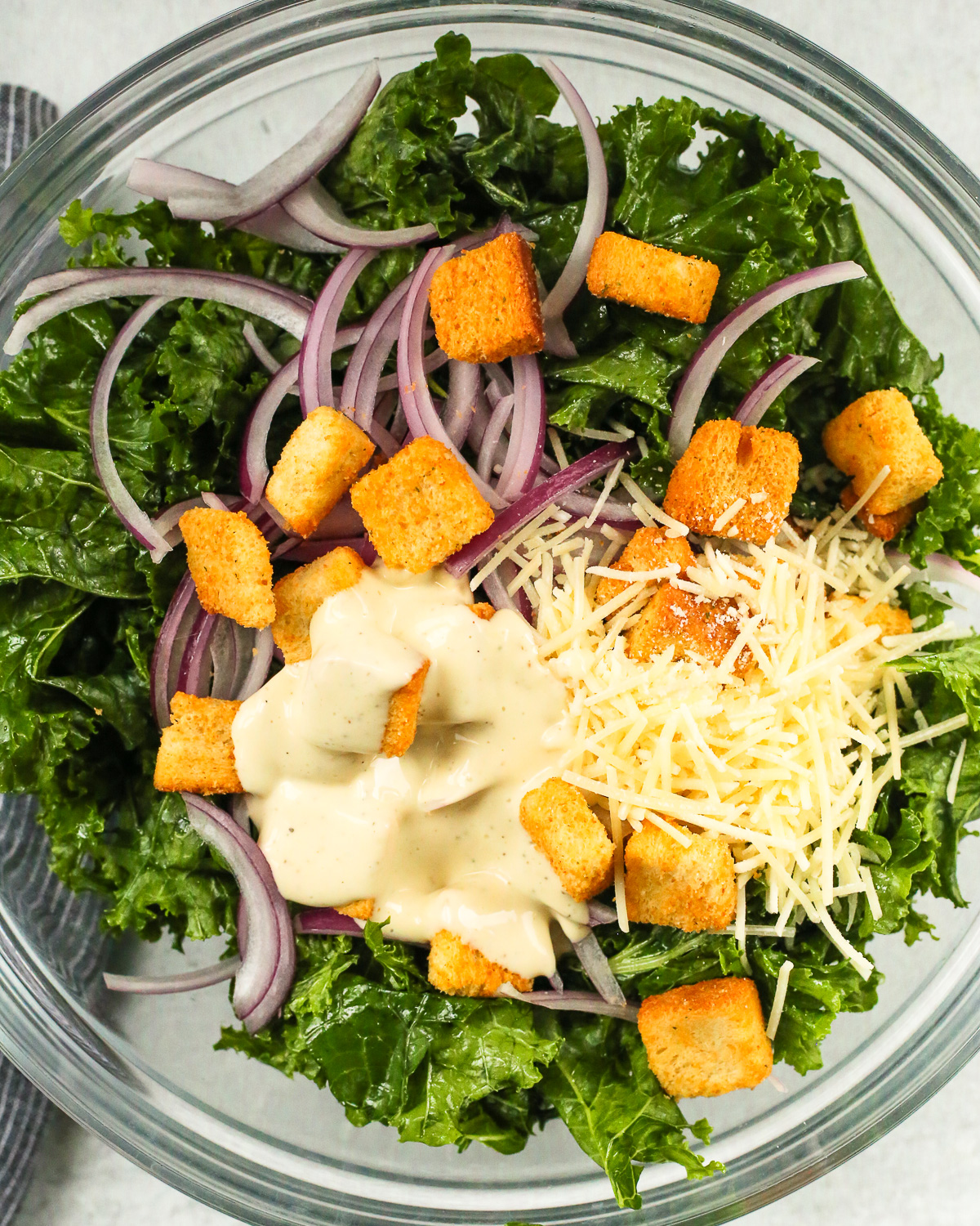Overhead view of a large mixing bowl, filled with massaged kale, caesar salad dressing, shredded parmesan cheese, sliced red onions, and crispy croutons that have not been mixed together yet.