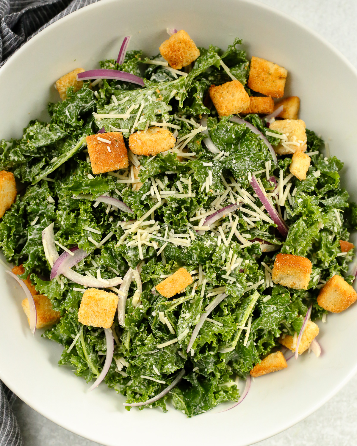 A white serving bowl filled with a massaged kale caesar salad, with sliced red onions and croutons mixed throughout.