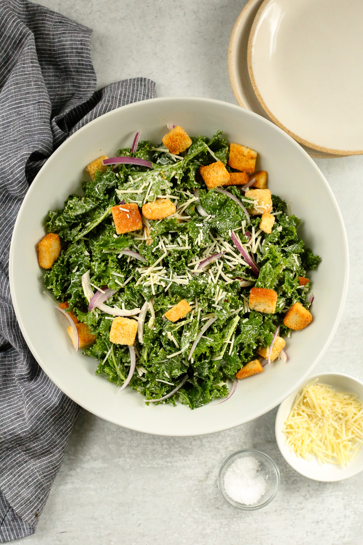 Overhead view of a large serving bowl containing a massaged kale caesar salad, topped with shredded parmesan cheese and golden brown croutons, with small salad plates arranged nearby as if preparing to serve the salad.