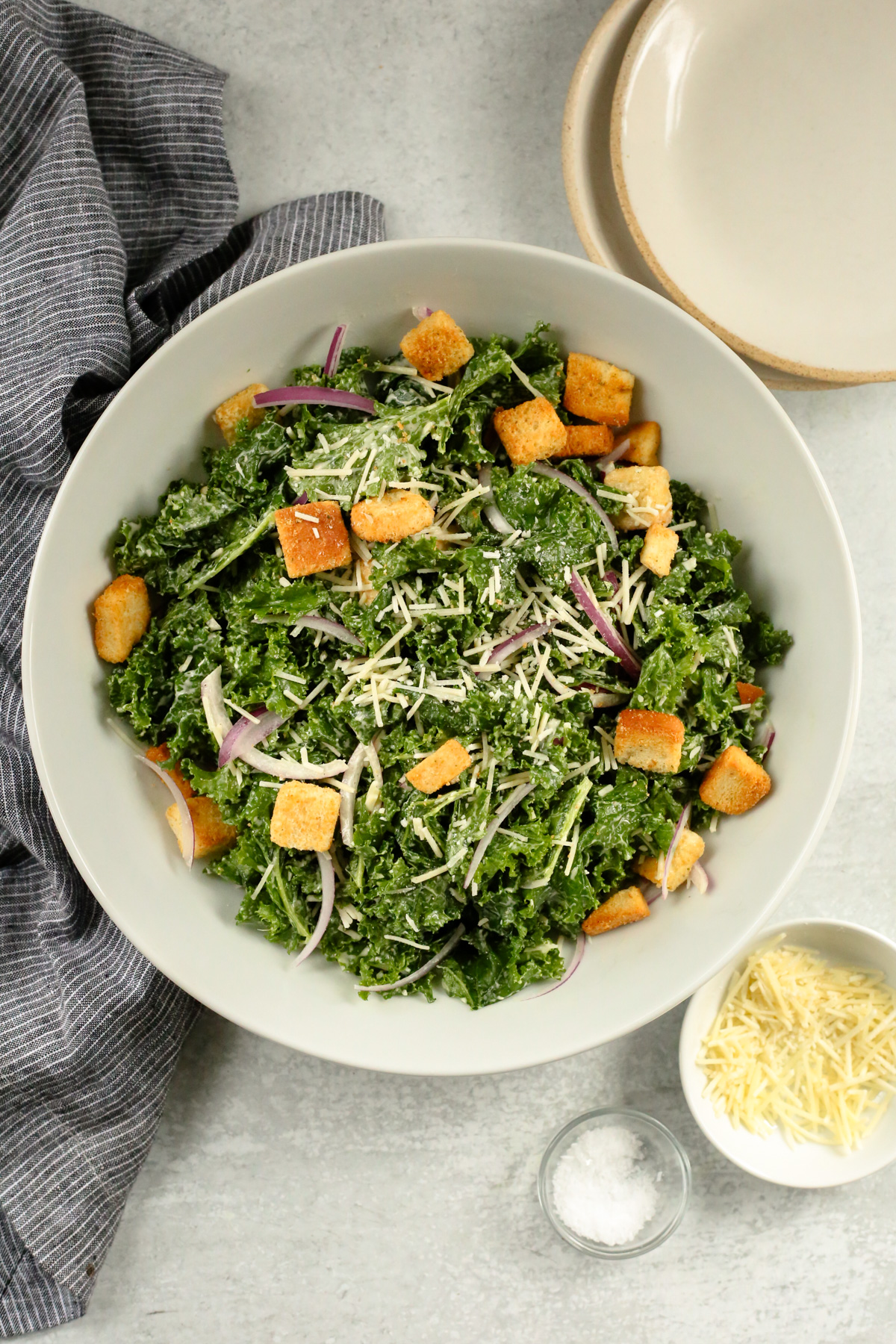 Overhead view of a large serving bowl containing a massaged kale caesar salad, topped with shredded parmesan cheese and golden brown croutons, with small salad plates arranged nearby as if preparing to serve the salad