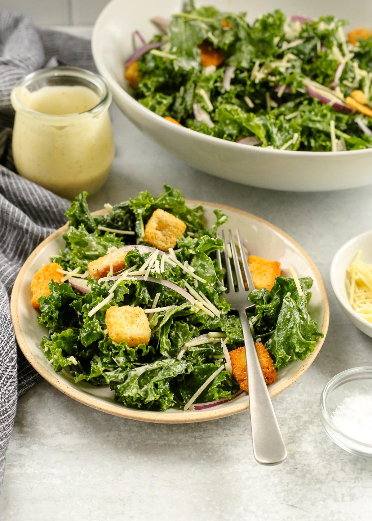 Angled view of a small ceramic salad plate piled high with massaged kale coated in Caesar salad dressing, with sliced red onions, parmesan cheese, and croutons mixed throughout, and a small glass jar of Caesar dressing and serving bowl with the remaining salad in the background.
