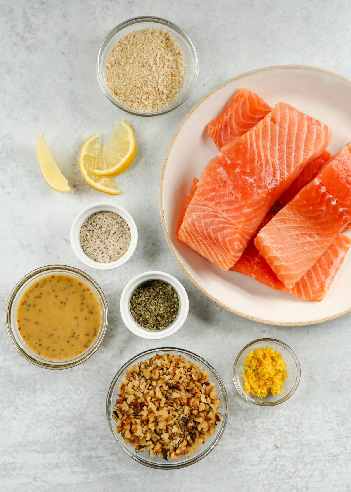 A display of the ingredients needed to make a walnut crusted salmon recipe, including uncooked salmon fillets, an all purpose seasoning blend, dried herbs, a maple dijon glaze, lemon zest, and chopped walnuts, arranged on a kitchen countertop