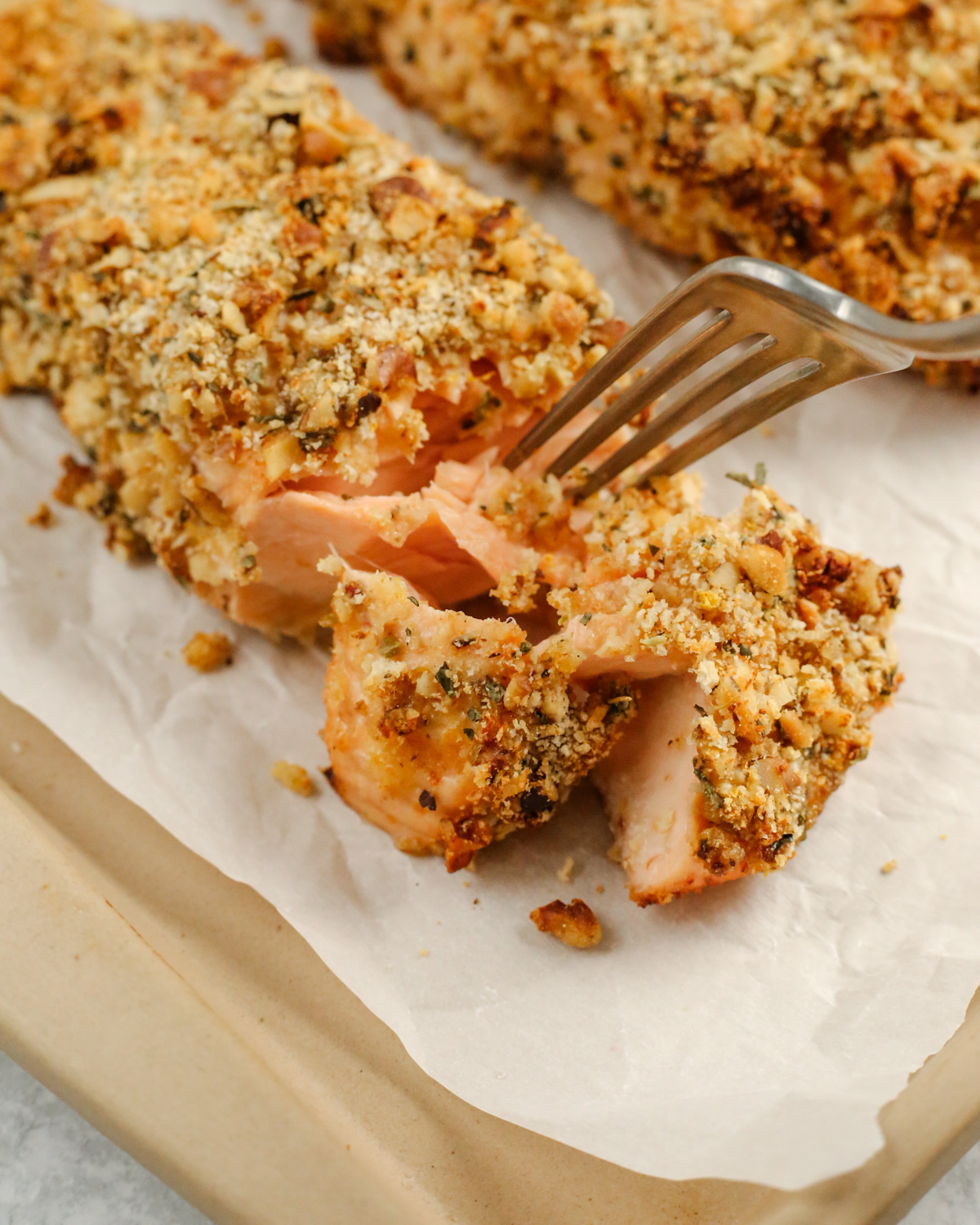 A silver fork prods a cooked fillet of salmon that is crusted with a walnut-breadcrumb mixture. The salmon flakes apart easily on the sheet pan, indicating it is perfectly cooked and ready to serve