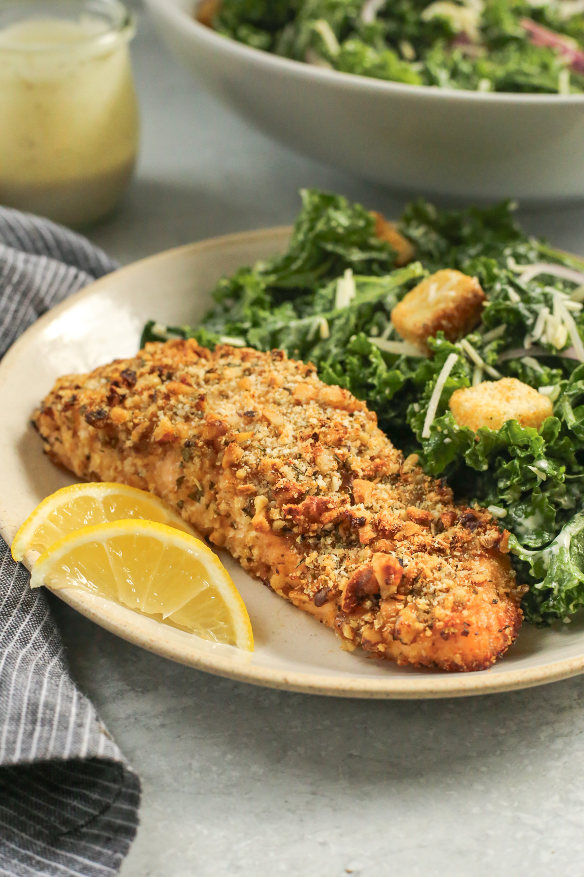 A serving of walnut crusted salmon on a ceramic plate, served with a kale caesar salad and croutons plus two small lemon wedges, with a serving bowl of salad in the background
