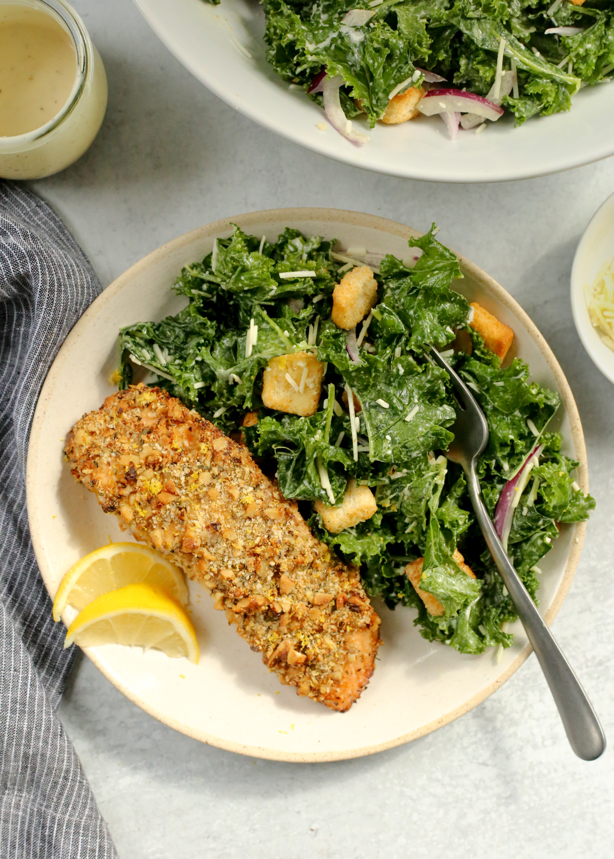 Overhead view of a cooked salmon fillet, coated in a walnut-breadcrumb crust, served with a side salad and lemon wedges with a silver fork arranged on the side of the dinner plate