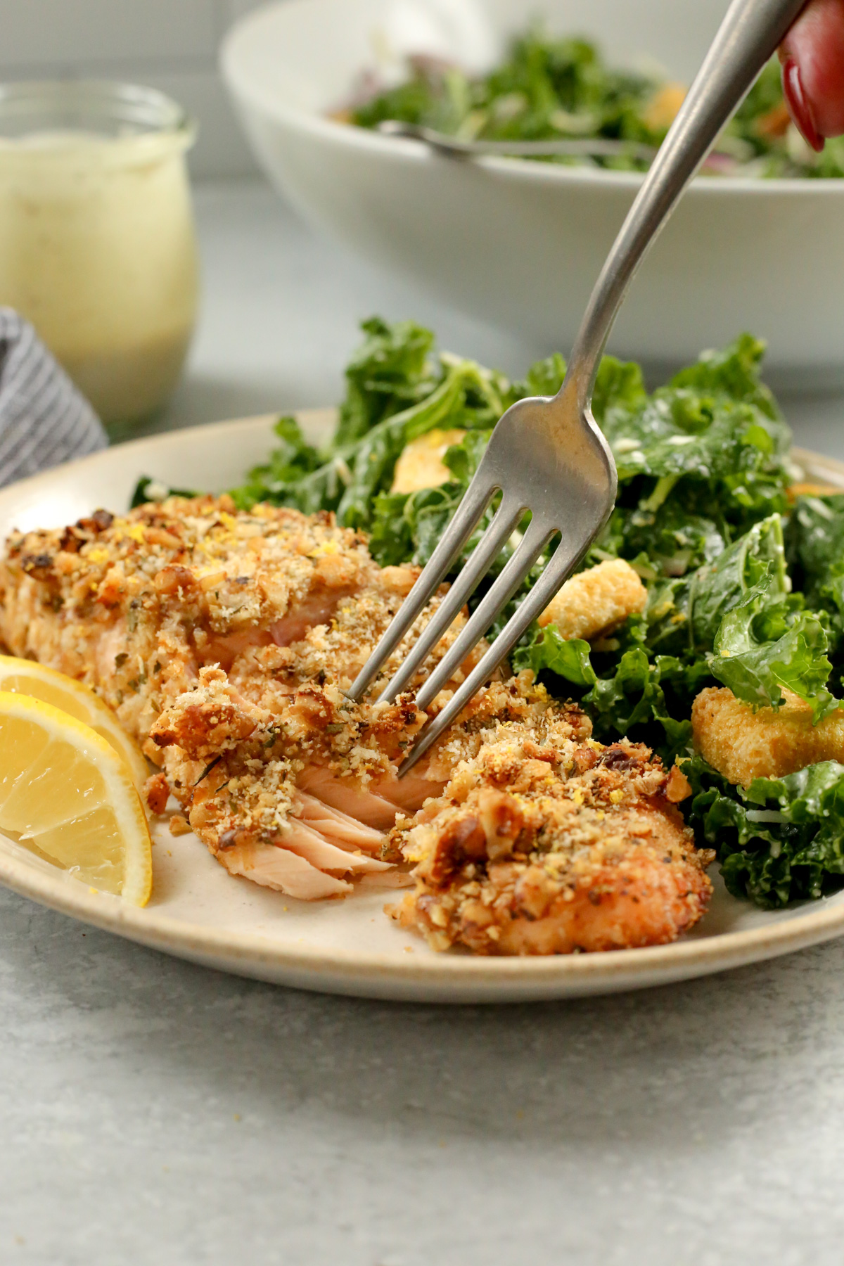 A silver fork reaches on a dinner plate to grab a bite of walnut crusted salmon, which is served with a side salad and two small lemon wedges