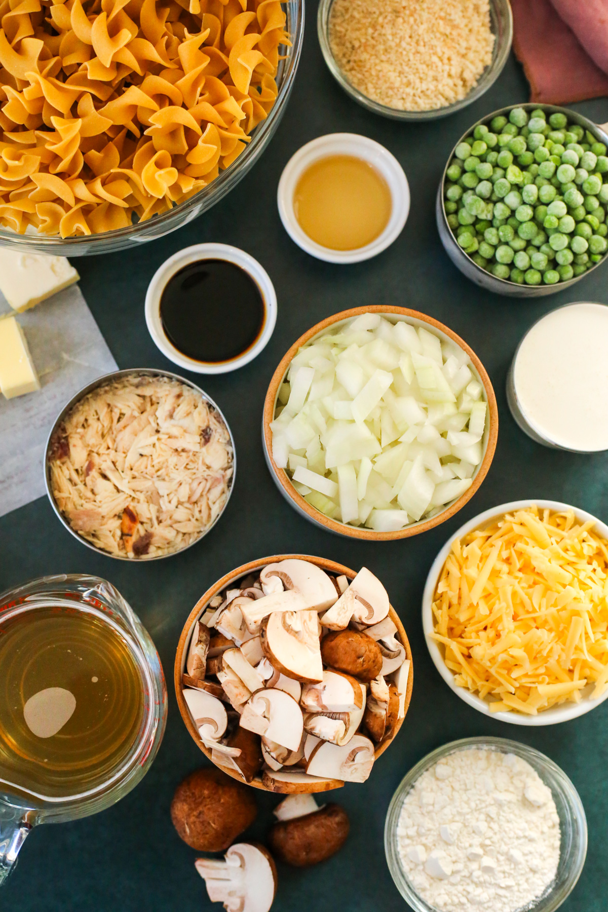 The ingredients needed to make a homemade tuna noodle casserole recipe are displayed on a dark teal background in various ramekins and prep bowls, including butter, egg noodles, panko bread crumbs, frozen green peas, diced onions, sliced mushrooms, soy sauce, apple cider vinegar, heavy cream, shredded cheese, all-purpose flour, and chicken broth