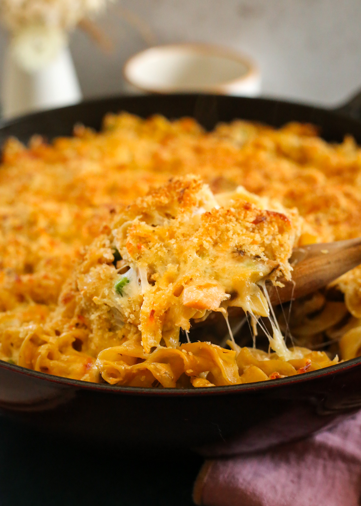 A wooden serving spoon lifts a heaping scoop of cheese tuna casserole from a dark red braising dish. The melted cheese stretches between the noodles and the crispy bread crump topping is golden brown