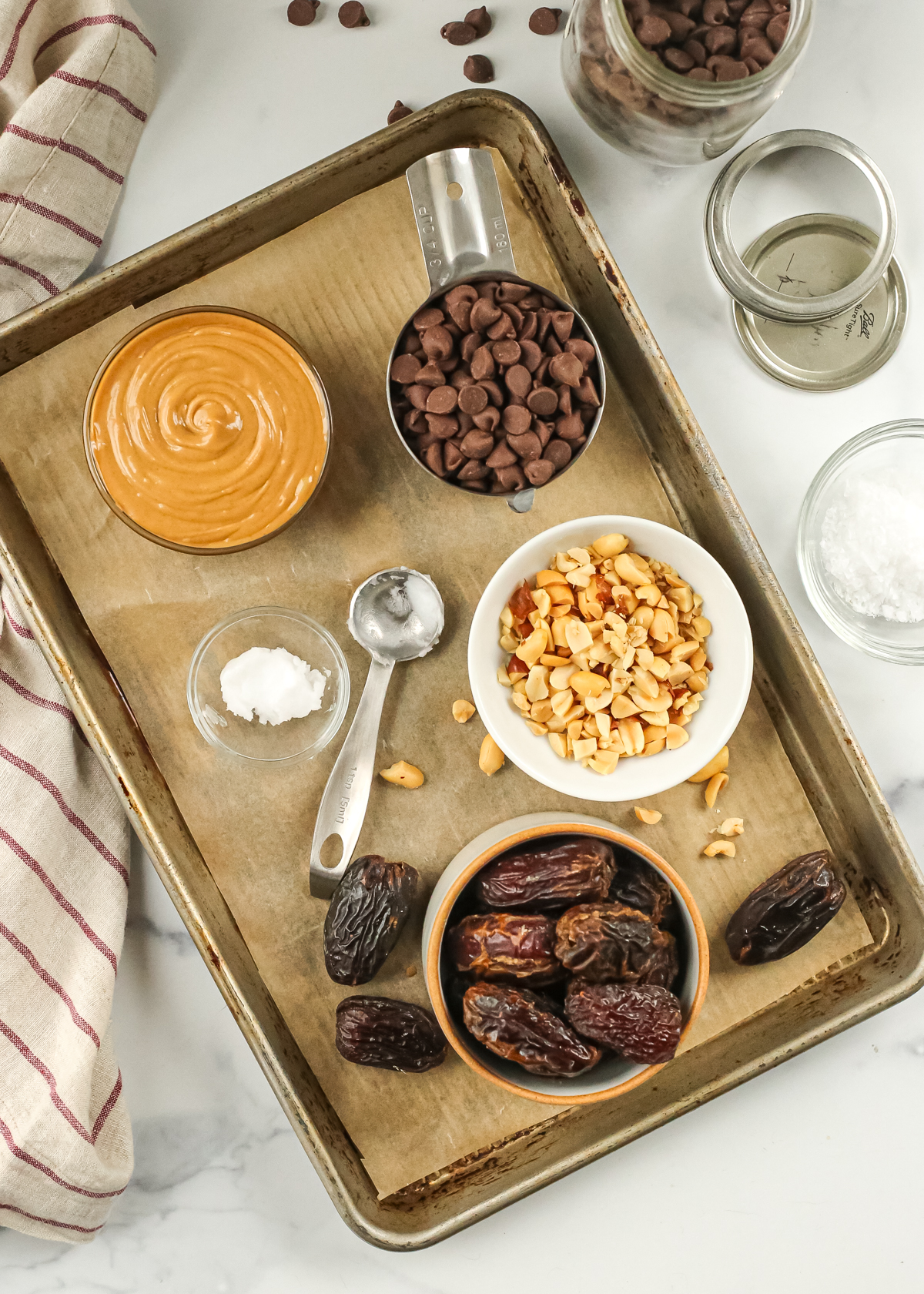 A baking tray is lined with a piece of parchment paper with the ingredients needed for date bark on top, including peanut butter, chocolate chips, roasted peanuts, coconut oil, and medjool dates