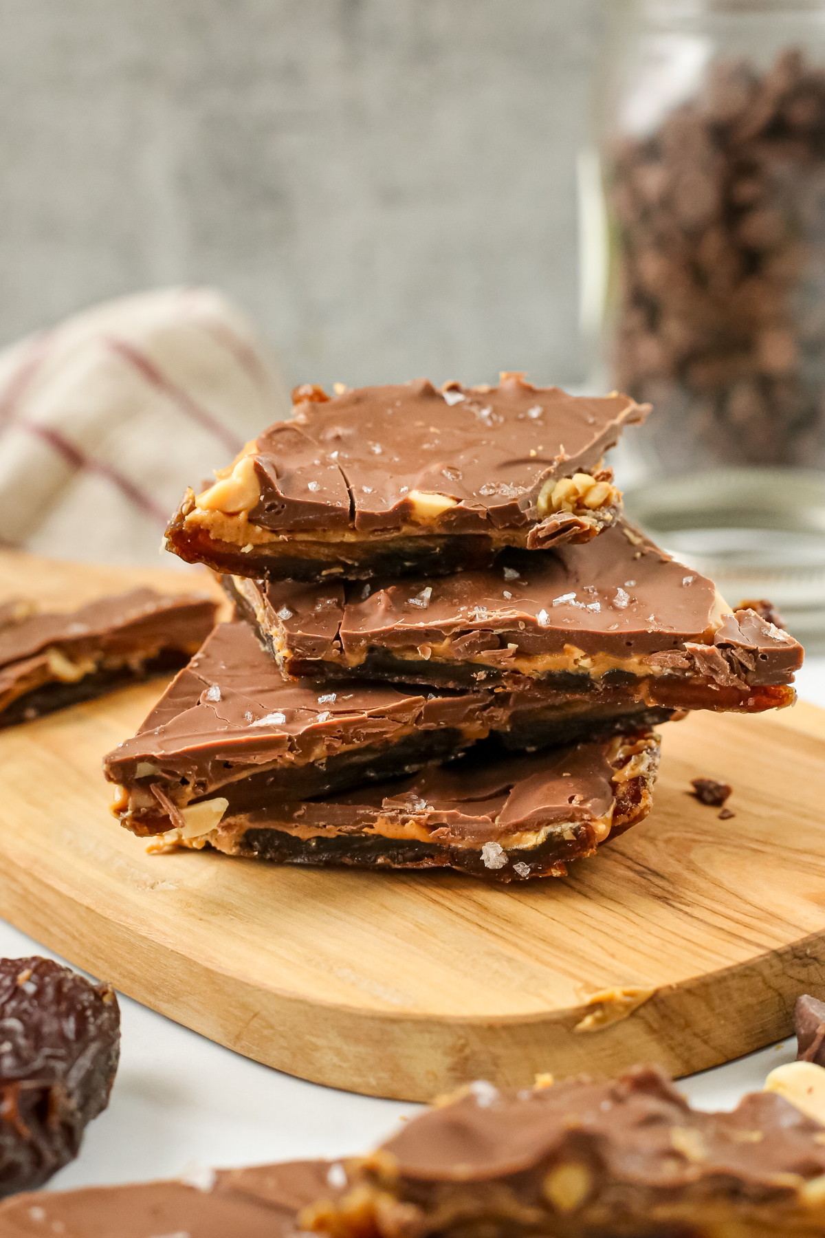 A stack of date bark is displayed on a small wooden serving board, showing layers of medjool dates, peanut butter, and chocolate topped with flaky sea salt