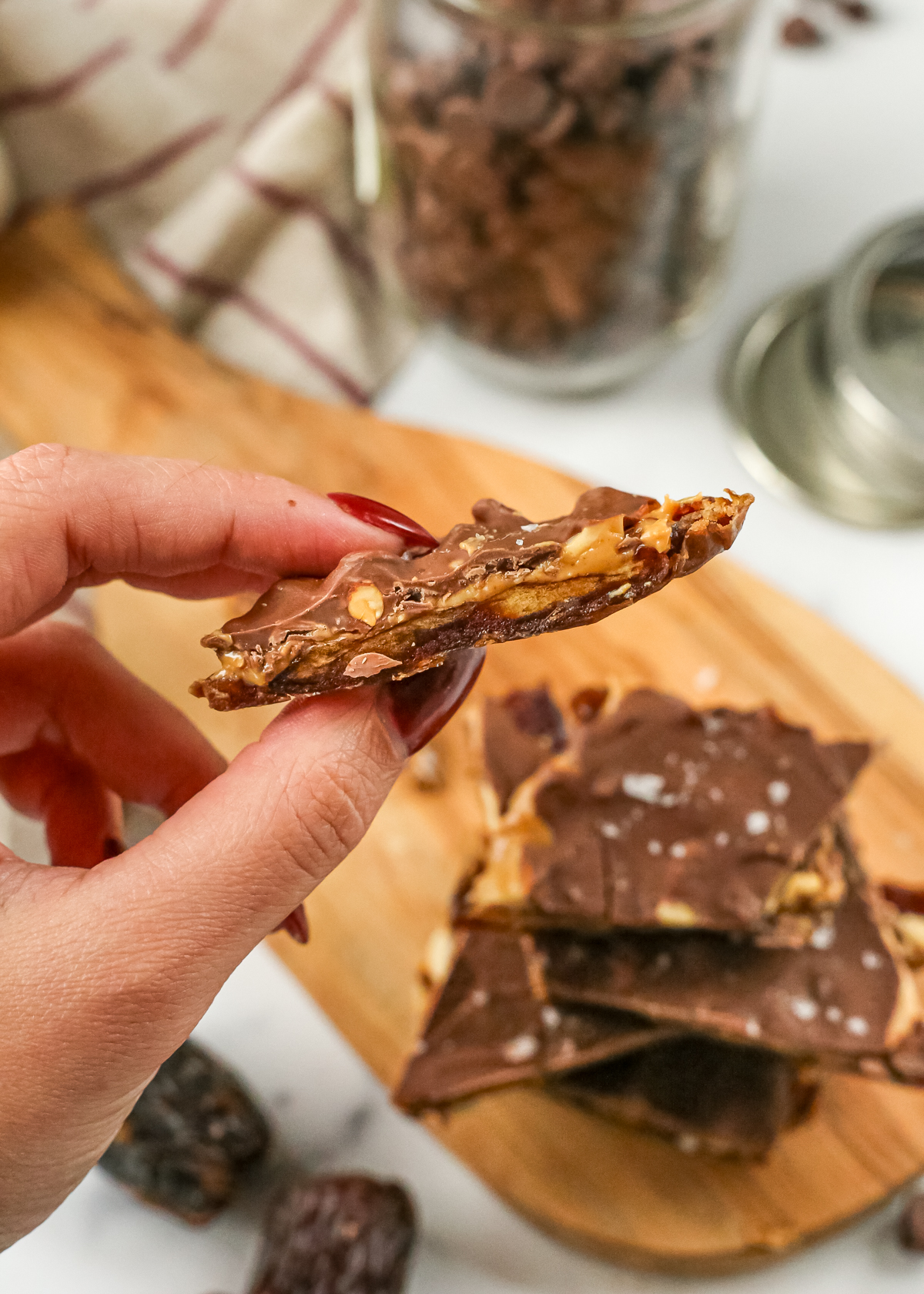 A woman's hand holds a piece of chocolate date bark, showing the layers of dates, peanut butter, and chocolate, with more pieces of date bark in the background in a kitchen setting