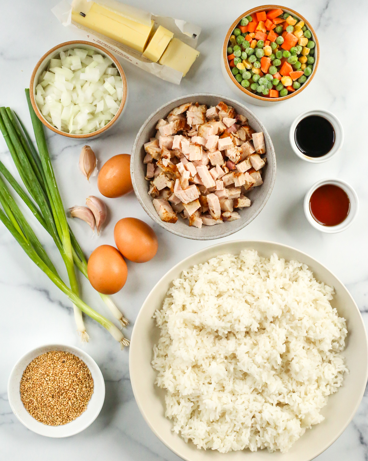 Overhead view of all the ingredients for this recipe, including cooked white rice, sesame seeds, eggs still in their brown shells, green onions, chopped white meat turkey, sesame oil, soy sauce, diced onions, garlic cloves, and a mixture of frozen veggies consisting of corn, peas, and carrots, and a stick of butter, all displayed in various sized prep bowls on a white marble kitchen countertop