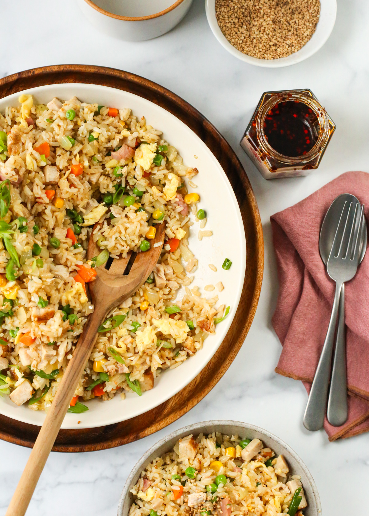 Overhead view of a serving bowl of turkey fried rice with a wooden serving spoon nestled on the right side, with a served portion in a smaller bowl in the lower portion of the image, and various garnishes displayed nearby