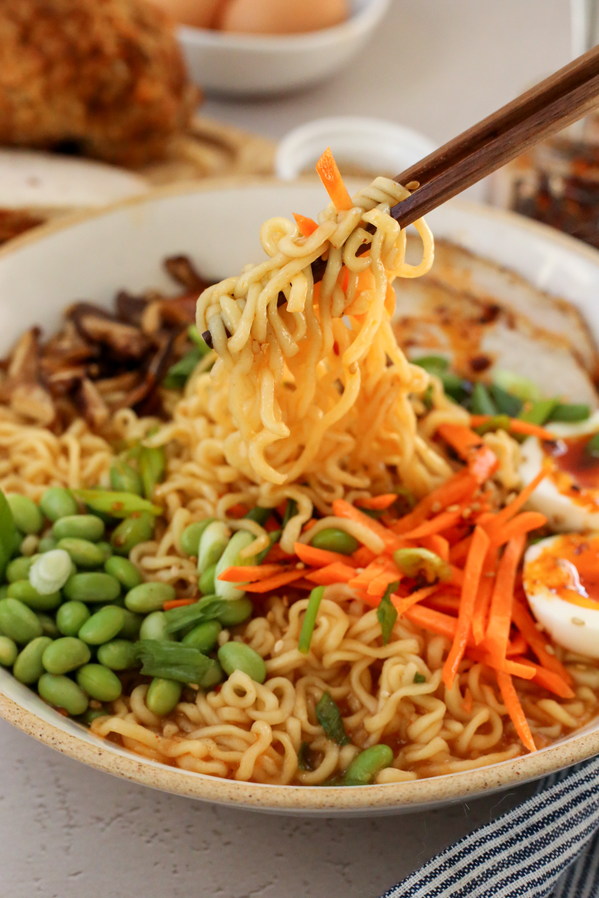 A pair of wooden chopsticks lifts a big bit of cooked ramen noodles out of a bowl filled with broth, with colorful vegetables mixed throughout