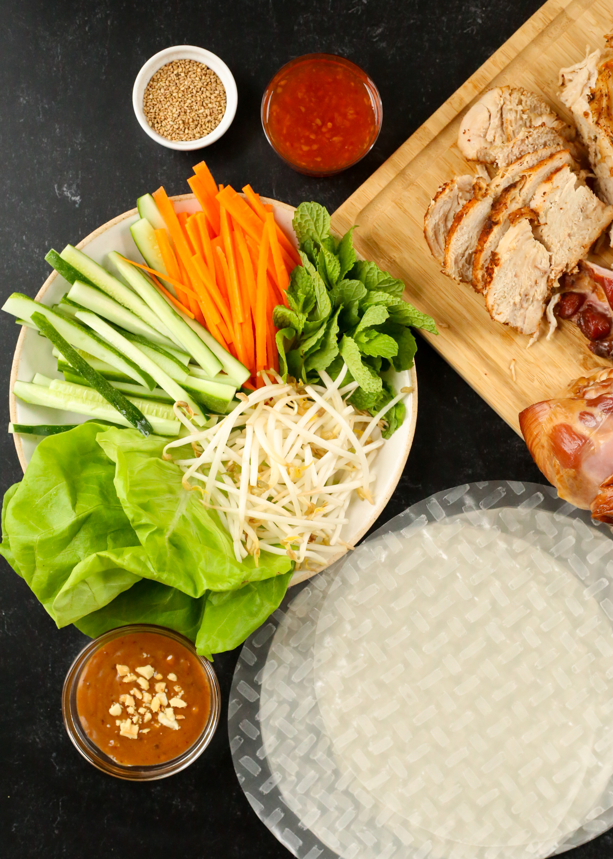 Overhead view of the ingredients needed to make a Leftover Turkey Spring Roll recipe, including a carving board with turkey, a plate full of thinly sliced carrots and cucumbers, lettuce leaves, bean sprouts, and mint leaves, round transparent rice paper wrappers stacked in the lower corner, and small prep bowls and ramekins with dipping sauces and toasted sesame seeds