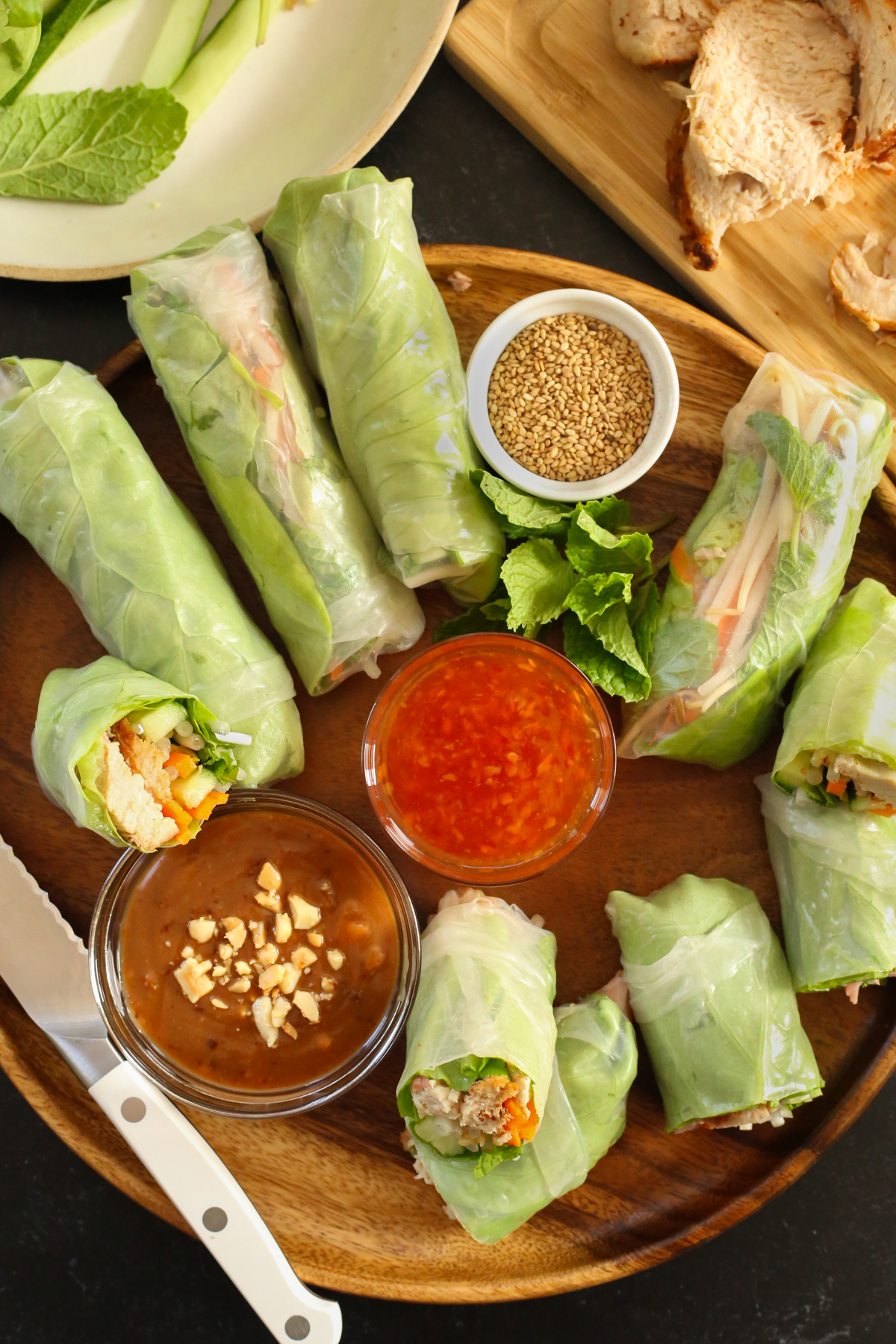 Overhead view of a tray full of spring rolls in various sizes. The tray is garnished with fresh mint leaves and there are two sauces served in small glass dishes to accompany the spring rolls, and a serrated knife with a white handle rests nearby to cut them in half