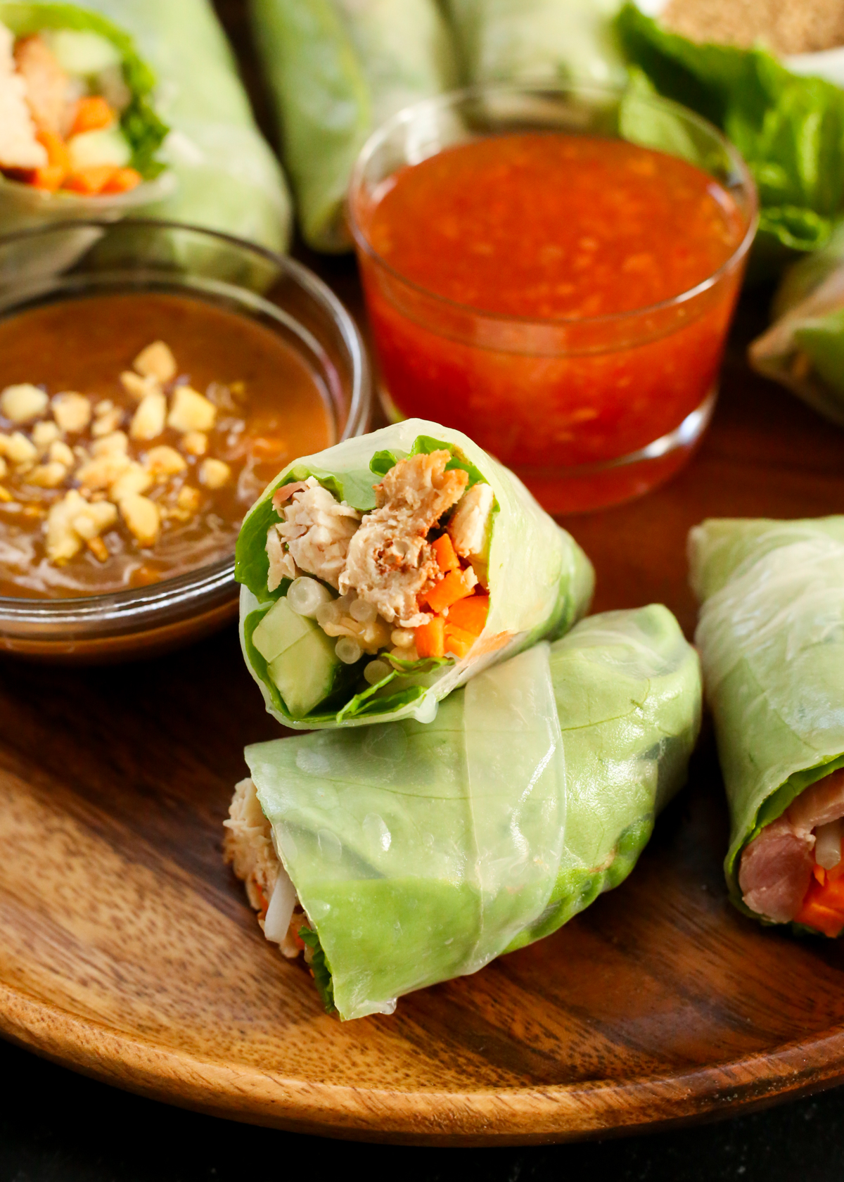 A cross sectional view into one of the turkey spring rolls, showing the shredded turkey meat, sliced cucumber and carrots, bean sprouts, mint leaves, and lettuce inside the spring roll wrapper. There are two different dipping sauces and additional spring rolls in the background