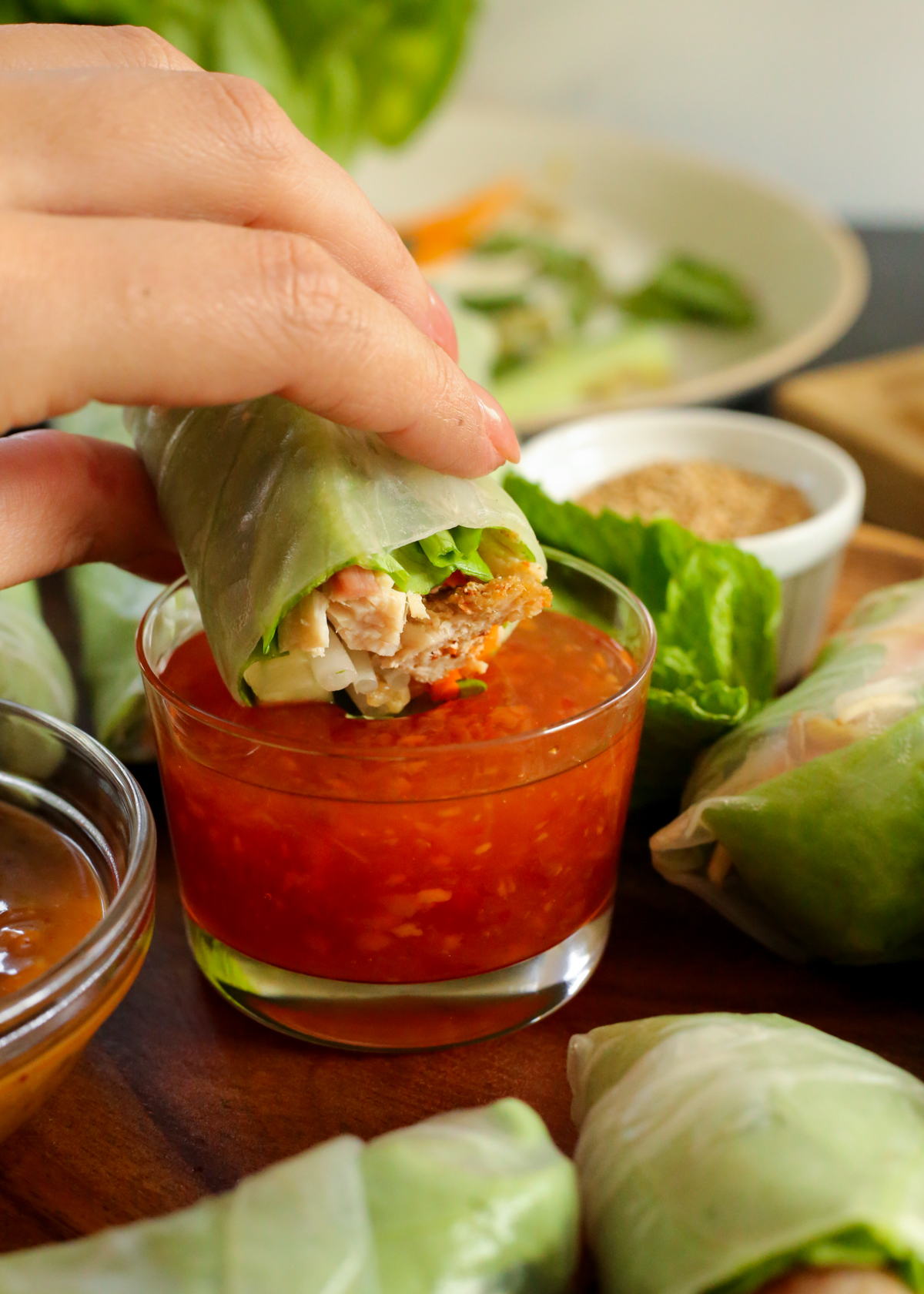 A woman's hand holds a turkey spring roll and dips it into a sweet chili sauce served in a small clear glass ramekin, with additional spring rolls and garnishes in the background