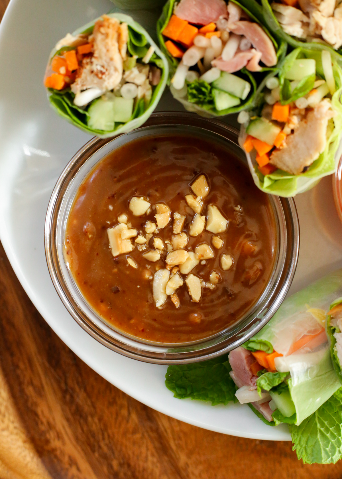 Overhead view of a peanut sauce topped with chopped peanuts, displayed on a serving platter filled with leftover turkey spring rolls that are tightly packed with turkey, carrots, cucumber, bean sprouts, mint leaves, and lettuce inside spring roll wrappers