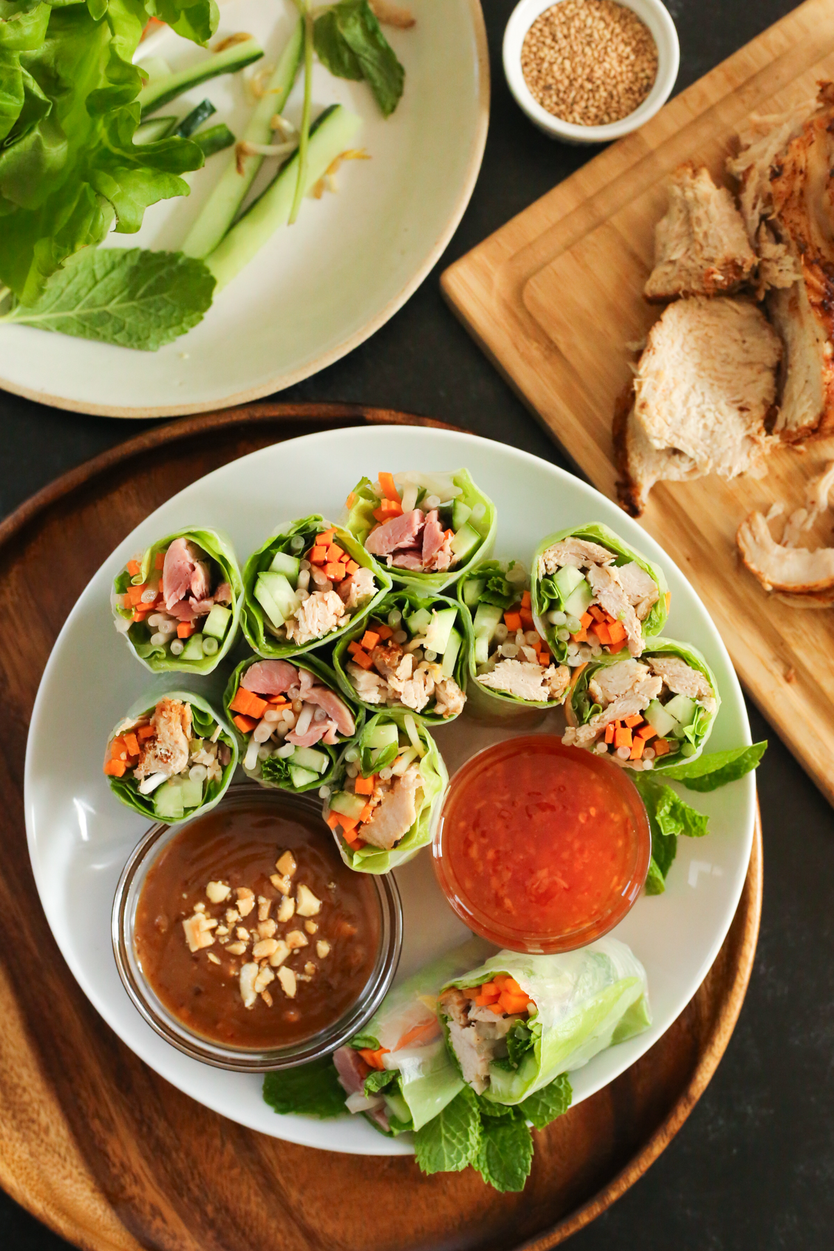 Overhead view of Leftover Turkey Spring Rolls arranged on a white plate and served on a dark wooden placemat, displayed with two different dipping sauces. The spring rolls are sliced through the middle to reveal carrots, turkey, cucumber, bean sprouts, and lettuce and mint leaves inside the rice paper wrappers