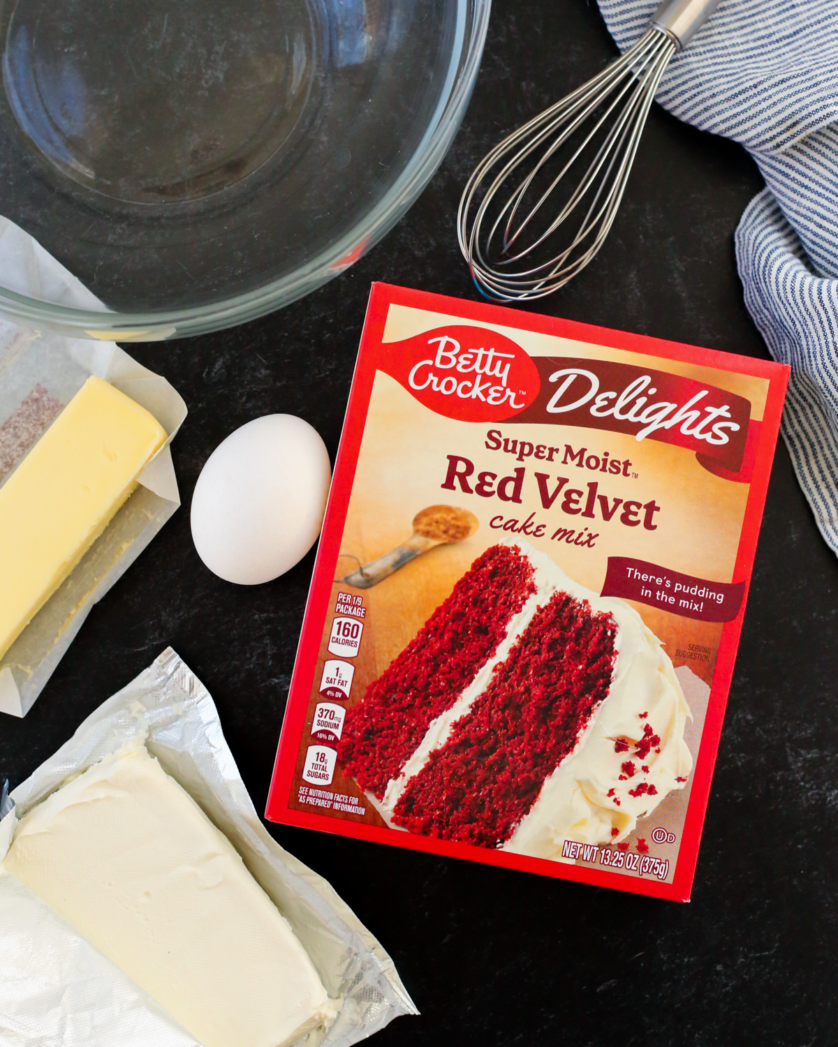 An overhead view of a box of Betty Crocker Super Moist Red Velvet Cake Mix is shown on a kitchen countertop next to an egg, a stick of butter, a block of cream cheese, a clear glass mixing bowl, and a stainless steel whisk