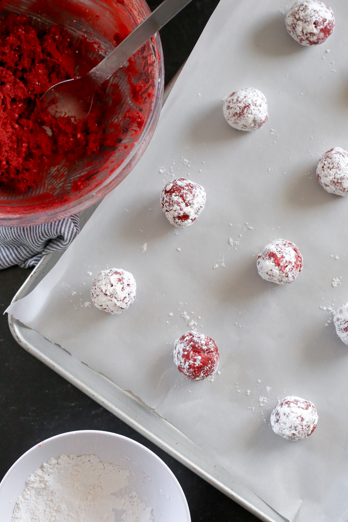 Overhead view of a large baking sheet lined with white parchment paper with prepared dough balls made using a red velvet cake mix and coated with powdered sugar