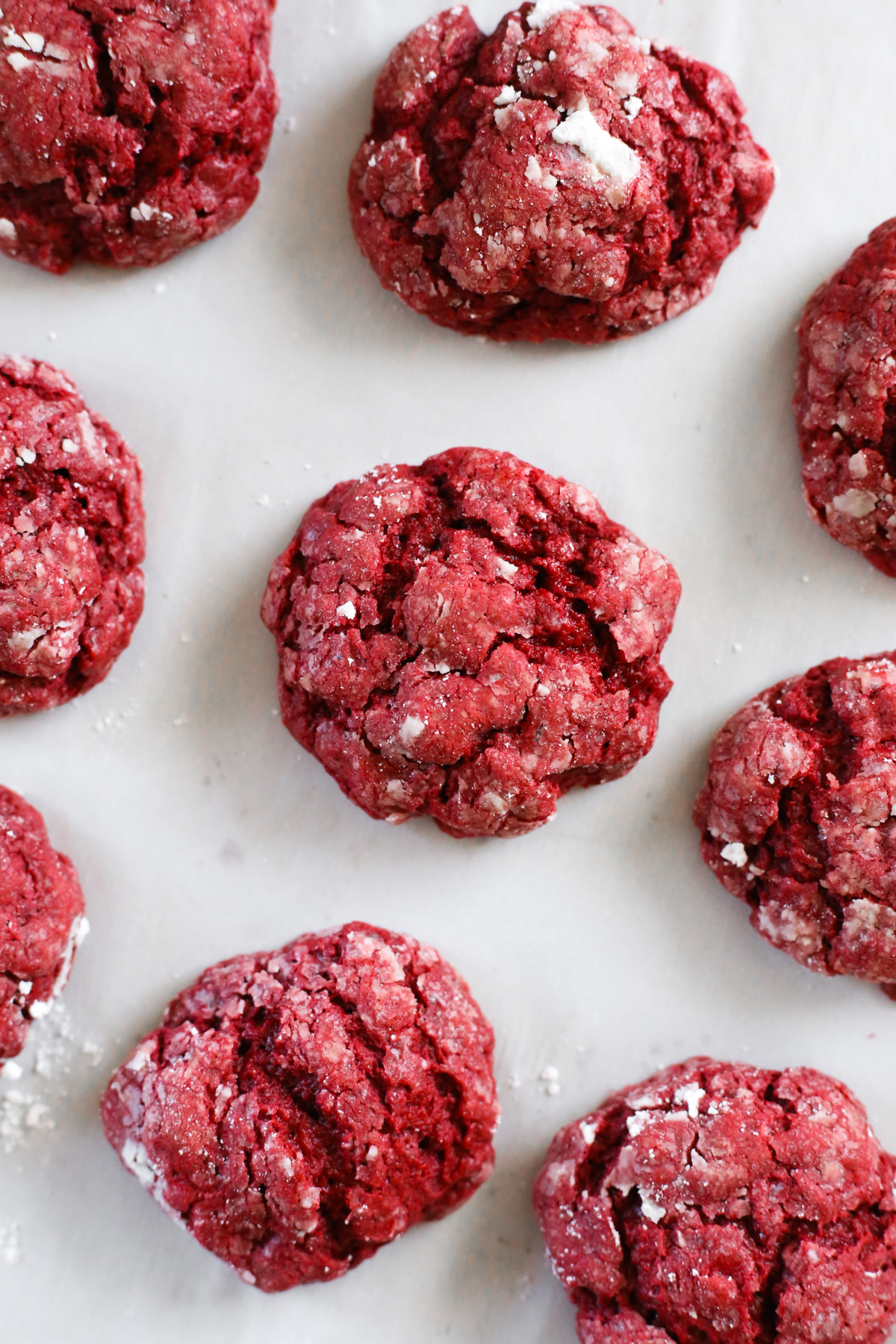 Bright red cookies with a cracked exterior and a dusting of powdered sugar are shown on a layer of white parchment paper as if they were freshly baked and just pulled from the oven