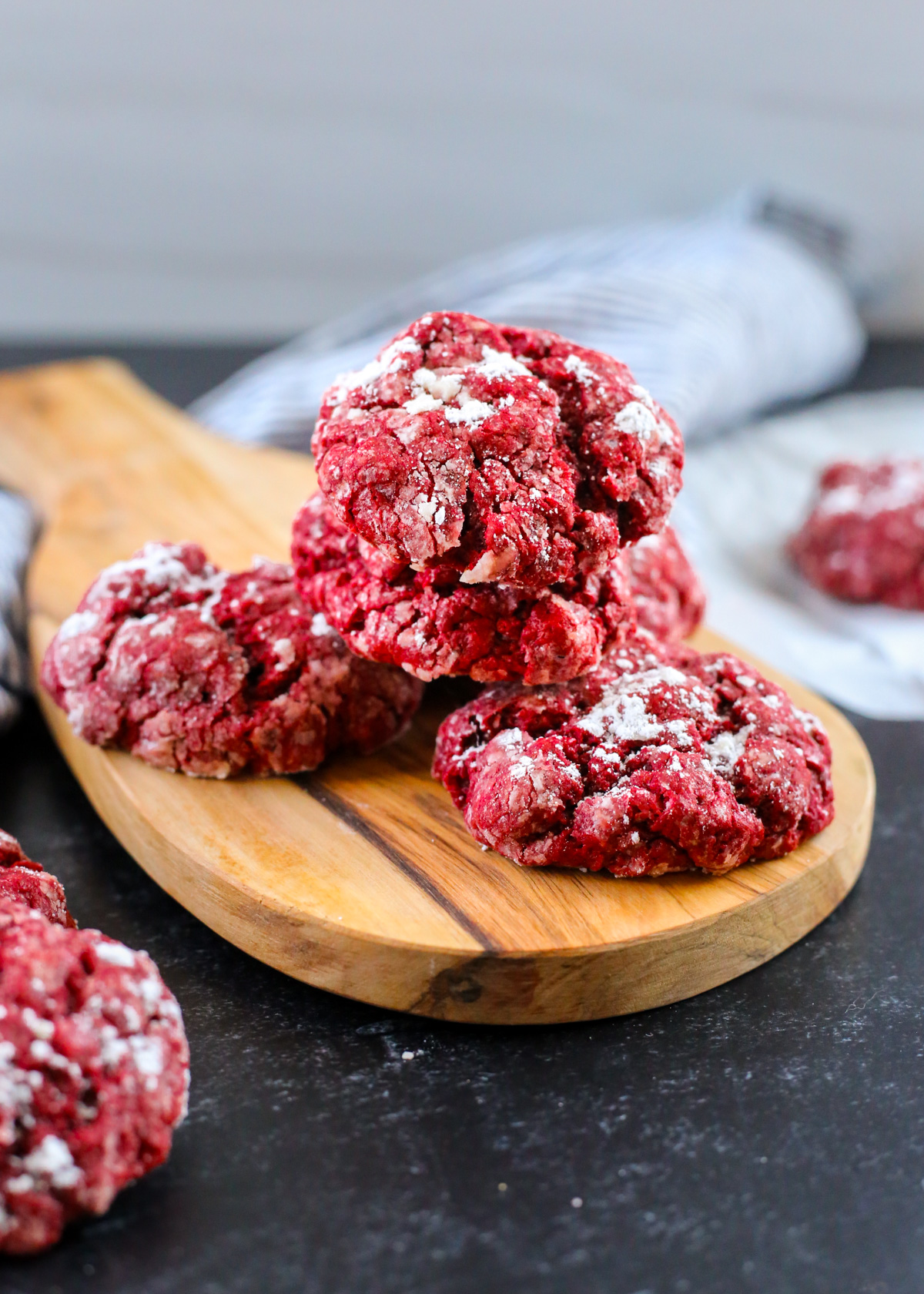 A stack of several red velvet gooey butter cookies is displayed on a small wooden serving board in a kitchen scene, with the cookies appearing moist and freshly baked with a light dusting on powdered sugar in contrast to the deep red color of the cookies